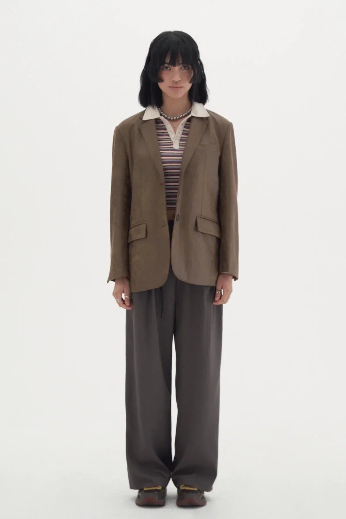 Model wearing brown blazer, striped polo shirt, wide gray slacks, and sneakers on white background