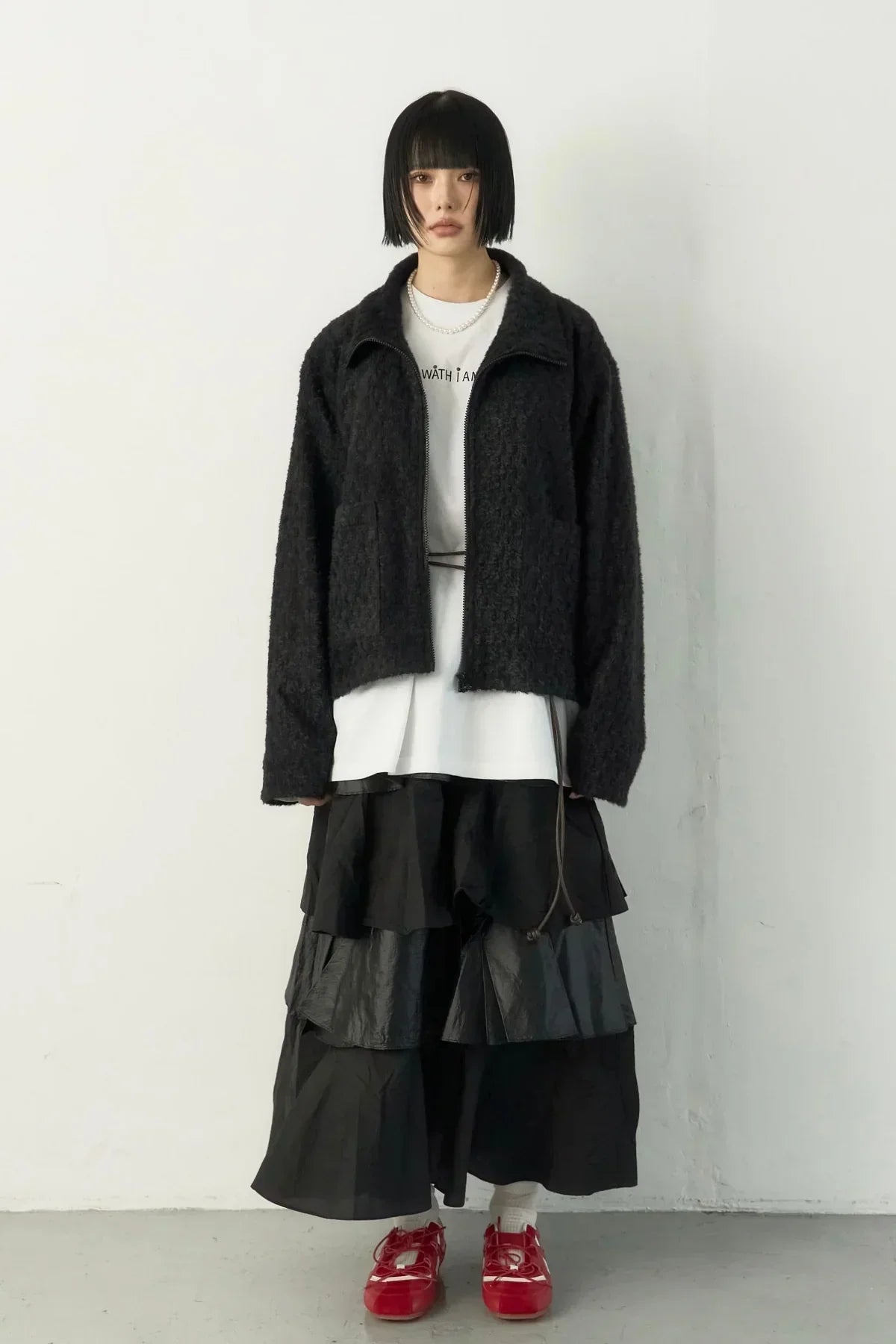 Model wearing black fuzzy zip jacket, white printed tee, layered black skirt, and red shoes in studio