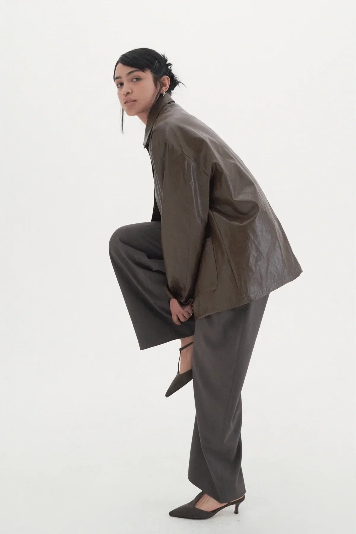 Model in dark wide slacks and brown oversized leather jacket posing against white background