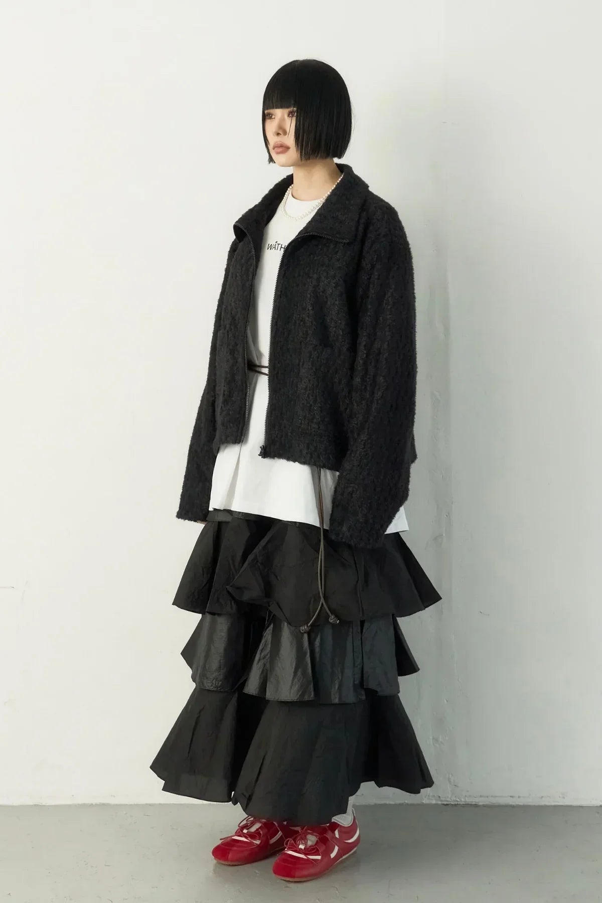 Woman with short black bob haircut wearing black textured jacket, white shirt, black layered skirt, and red sneakers