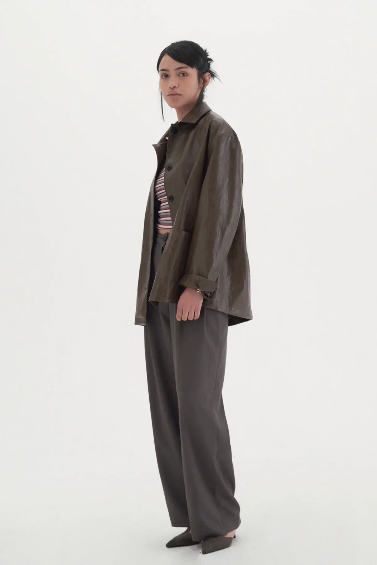 Woman in wide-leg gray slacks, brown oversized jacket, striped crop top, and pointed shoes against white background