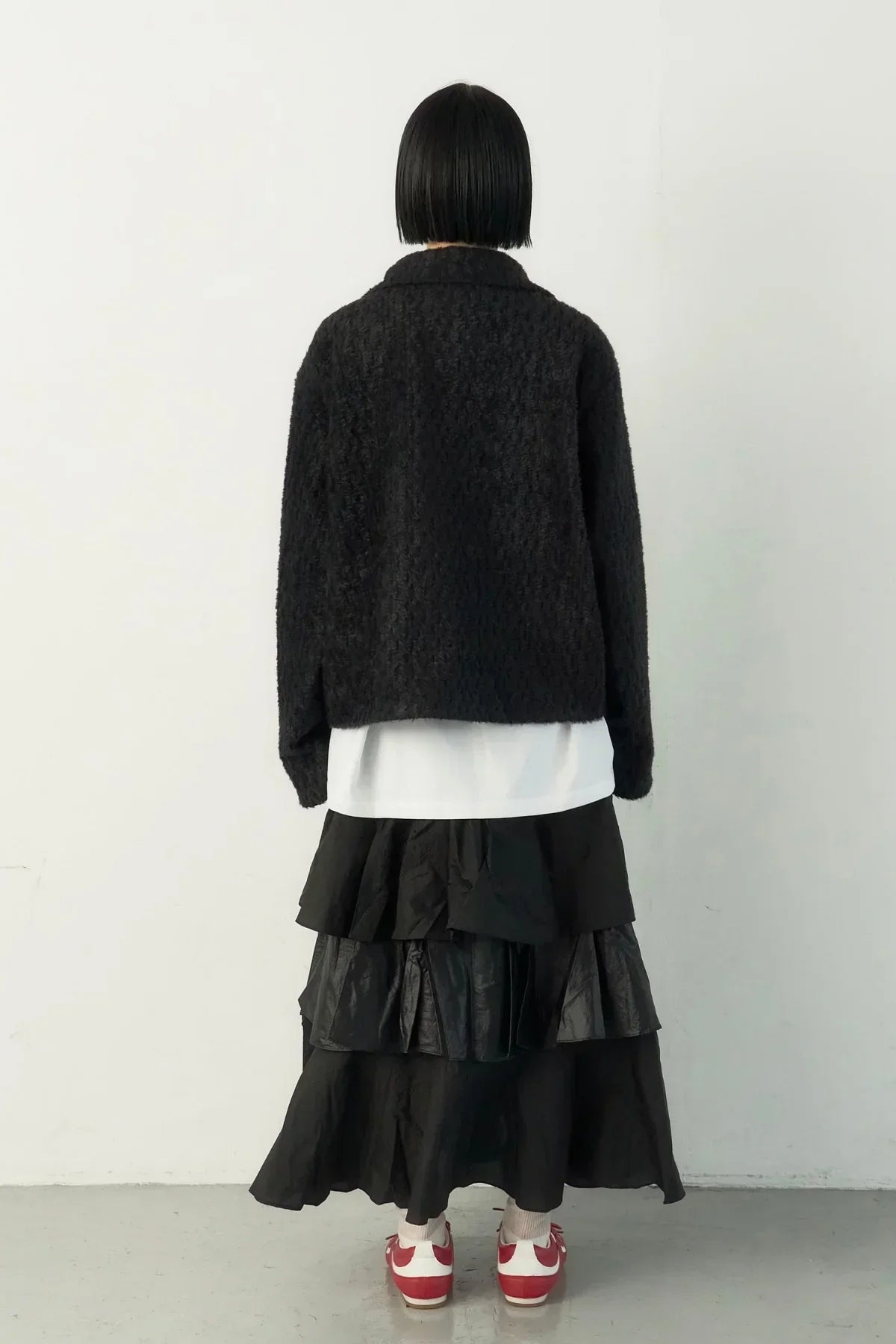 Person with short black hair wearing black fuzzy jacket, white shirt, tiered black skirt, red and white sneakers