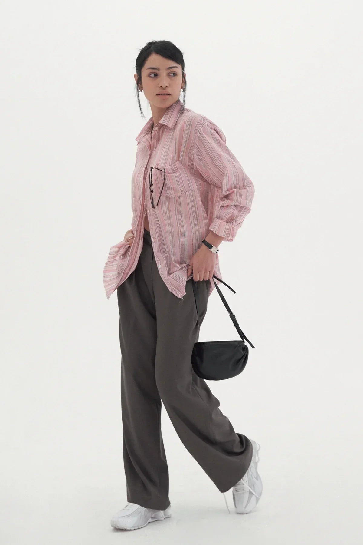 Woman in pink striped shirt, wide dark tuck slacks, white sneakers holding black handbag on white background