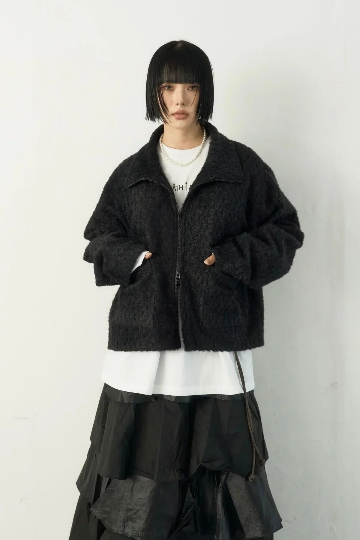 Model with black bob haircut wearing a textured black zip jacket, white shirt, layered black skirt, and pearl necklace