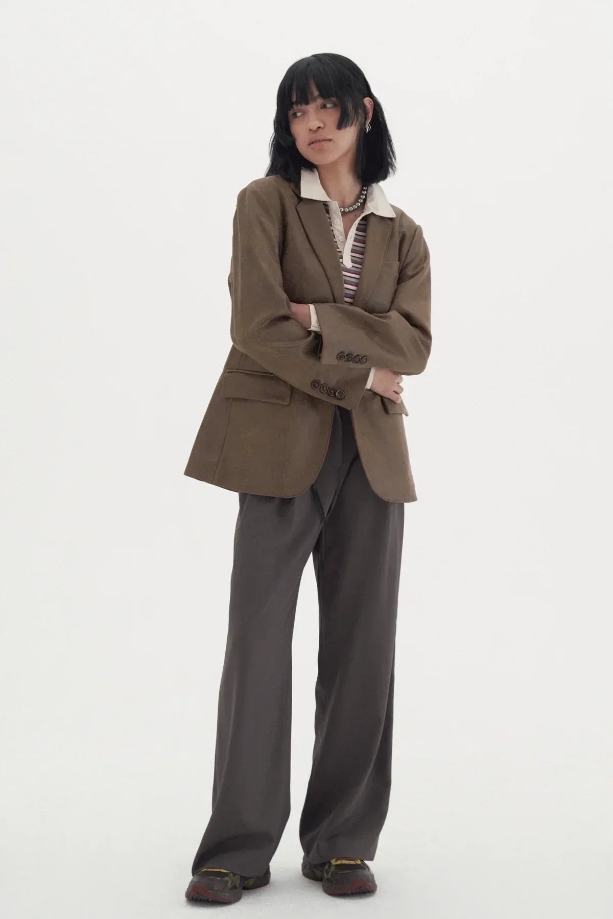 Model in brown blazer and wide dark slacks with striped shirt and sneakers on white background