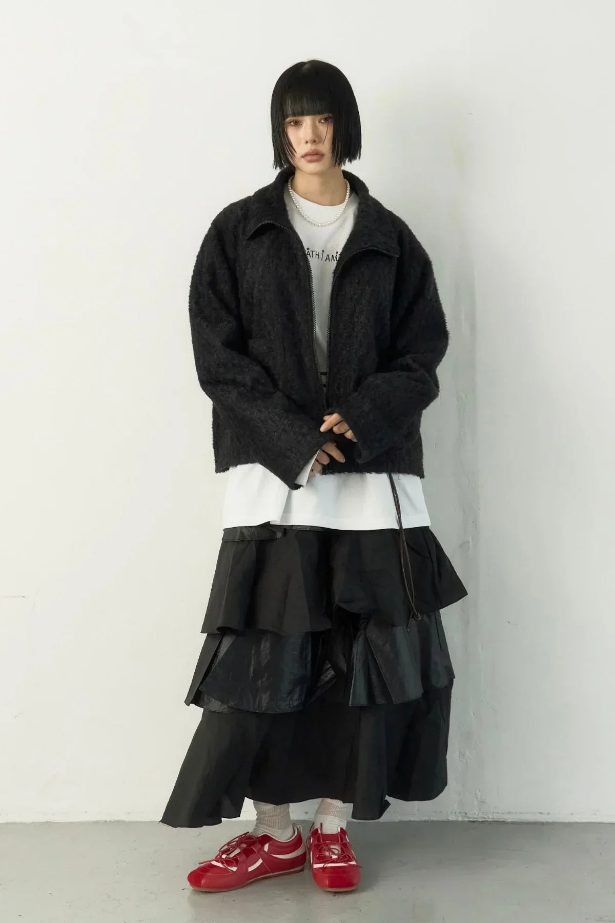 Woman wearing black fuzzy jacket, layered black skirt, white shirt, and red sneakers against white wall