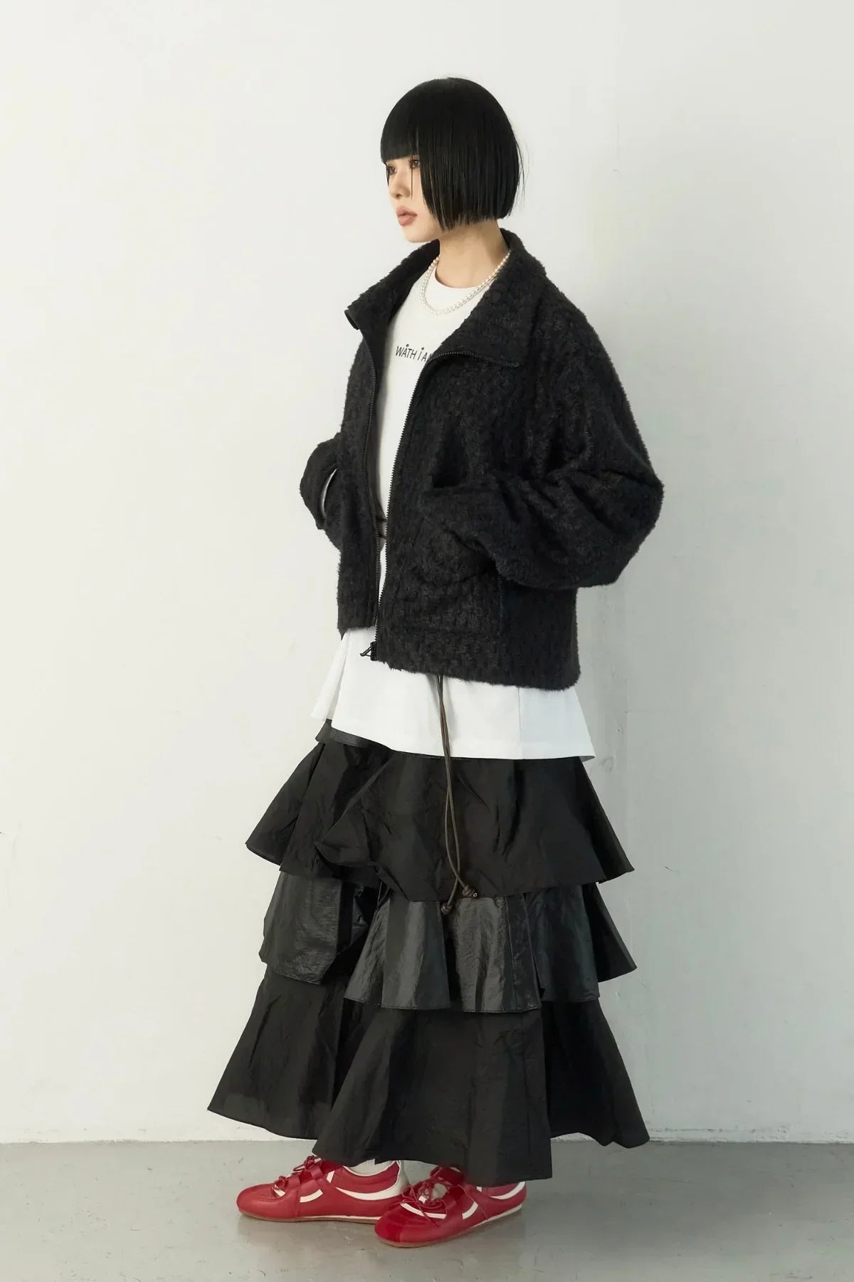 Woman with short bob haircut wearing a black textured zip-up jacket, layered black ruffled skirt, white shirt, and red sneakers
