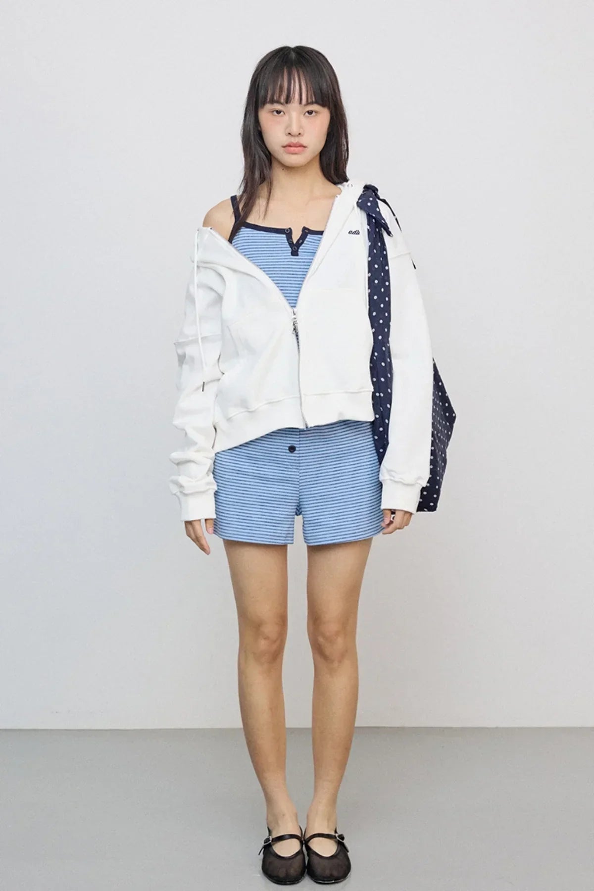 Young woman wearing an ivory zip-up hoodie, blue striped tank top and shorts, black flats, with a navy polka dot bag