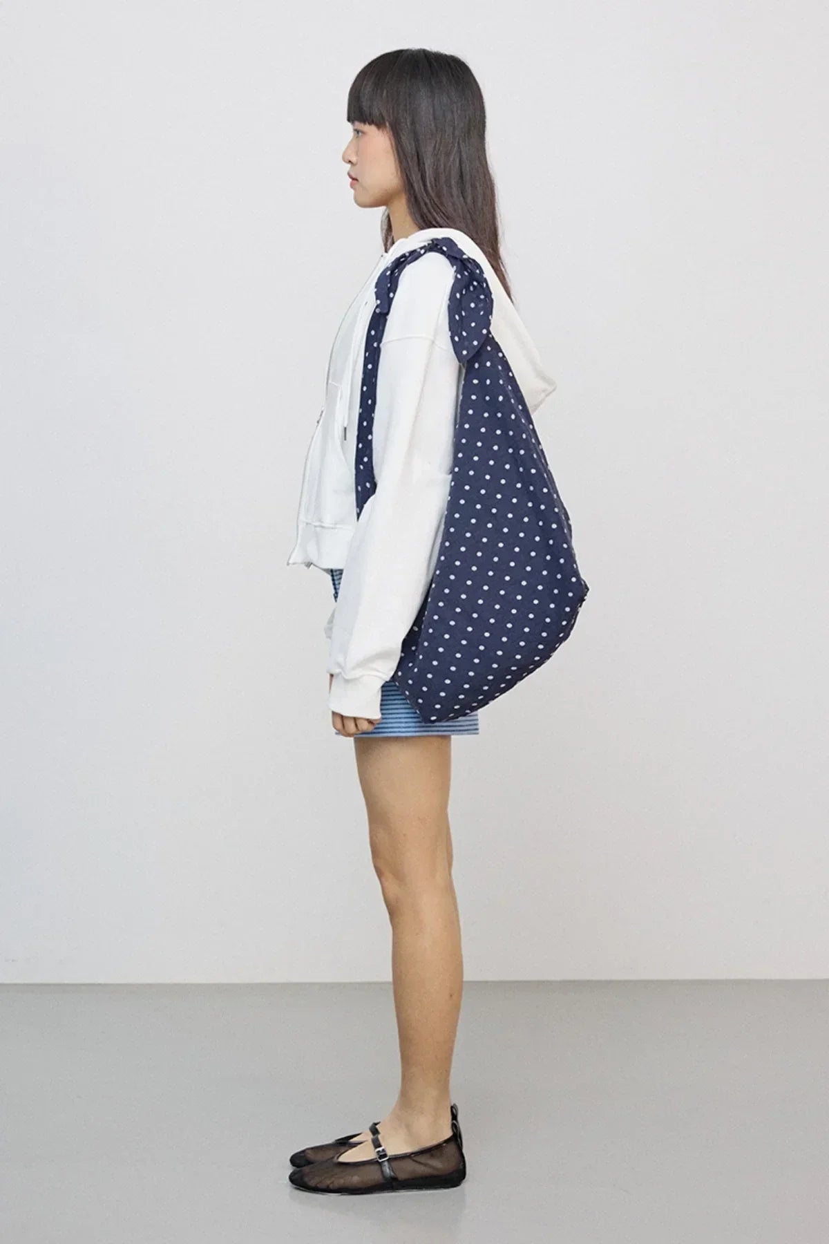 Side view of woman wearing ivory hoodie, striped shorts, black flats, carrying navy polka dot tote bag