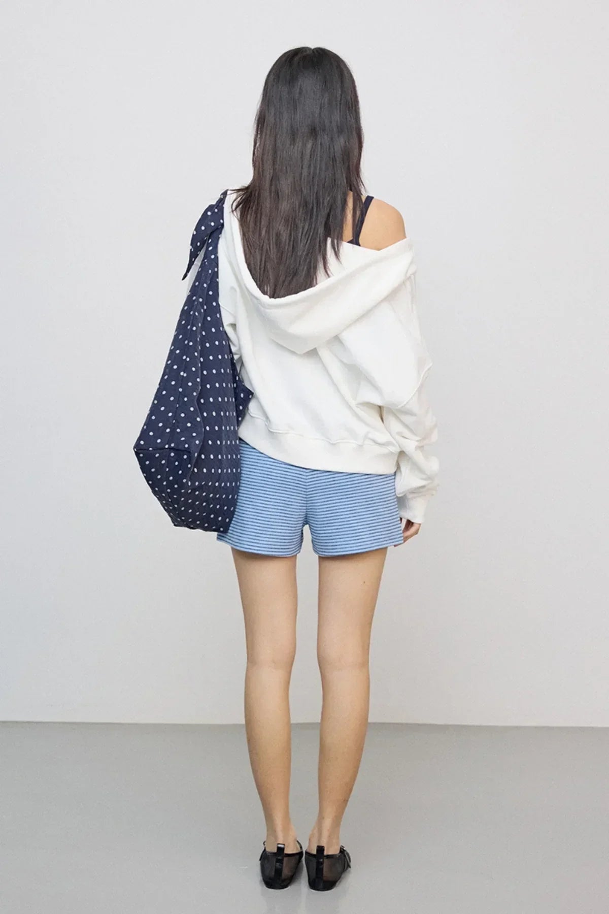 Woman with long dark hair wearing oversized off-white hoodie, blue striped shorts, carrying navy polka dot bag