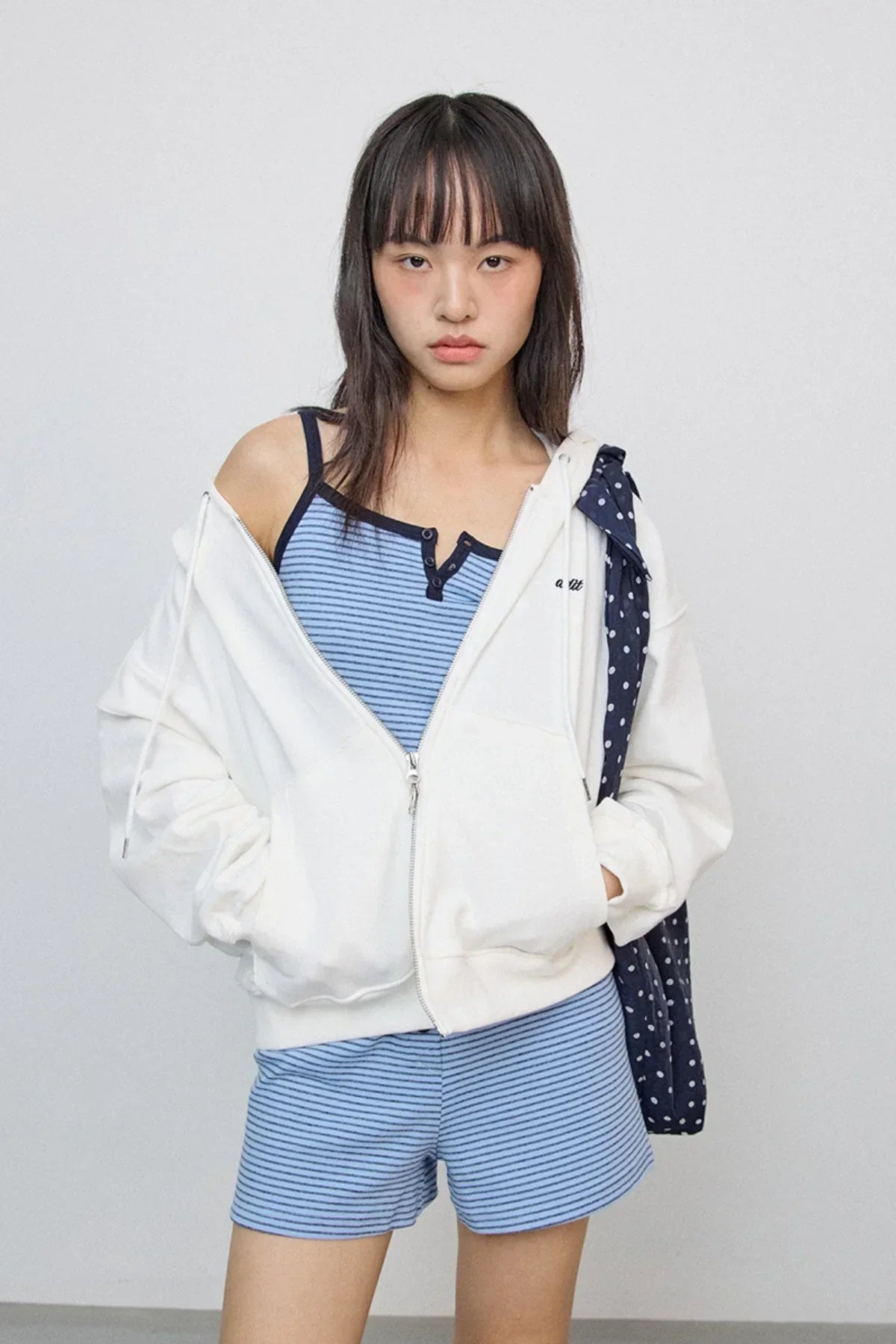 Young woman wearing ivory zip-up hoodie over blue striped tank top and shorts with polka dot bag