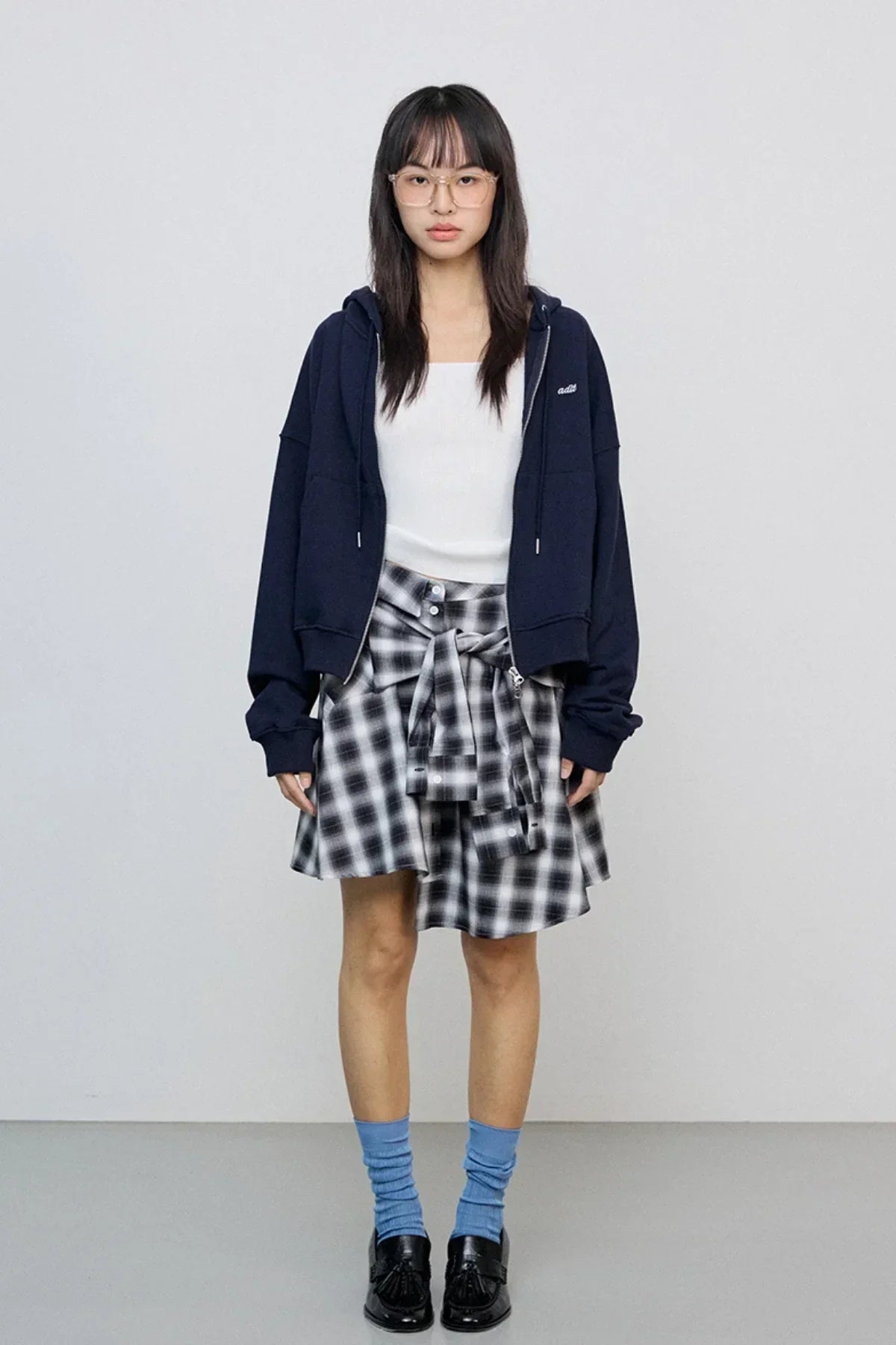 Woman wearing navy zip-up hoodie, white tank top, black plaid skirt, blue socks, and black shoes