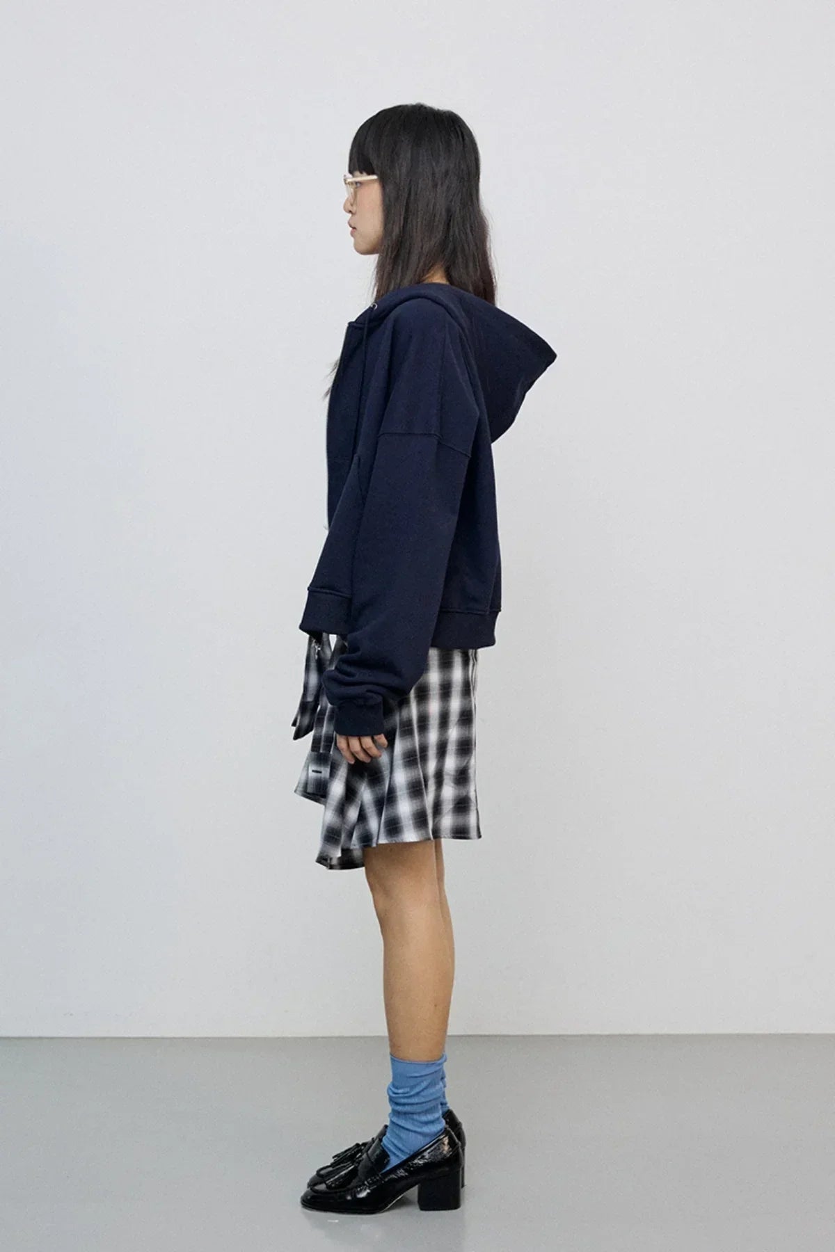 Side view of woman wearing navy zip-up hoodie, plaid skirt, blue socks, and black heeled loafers