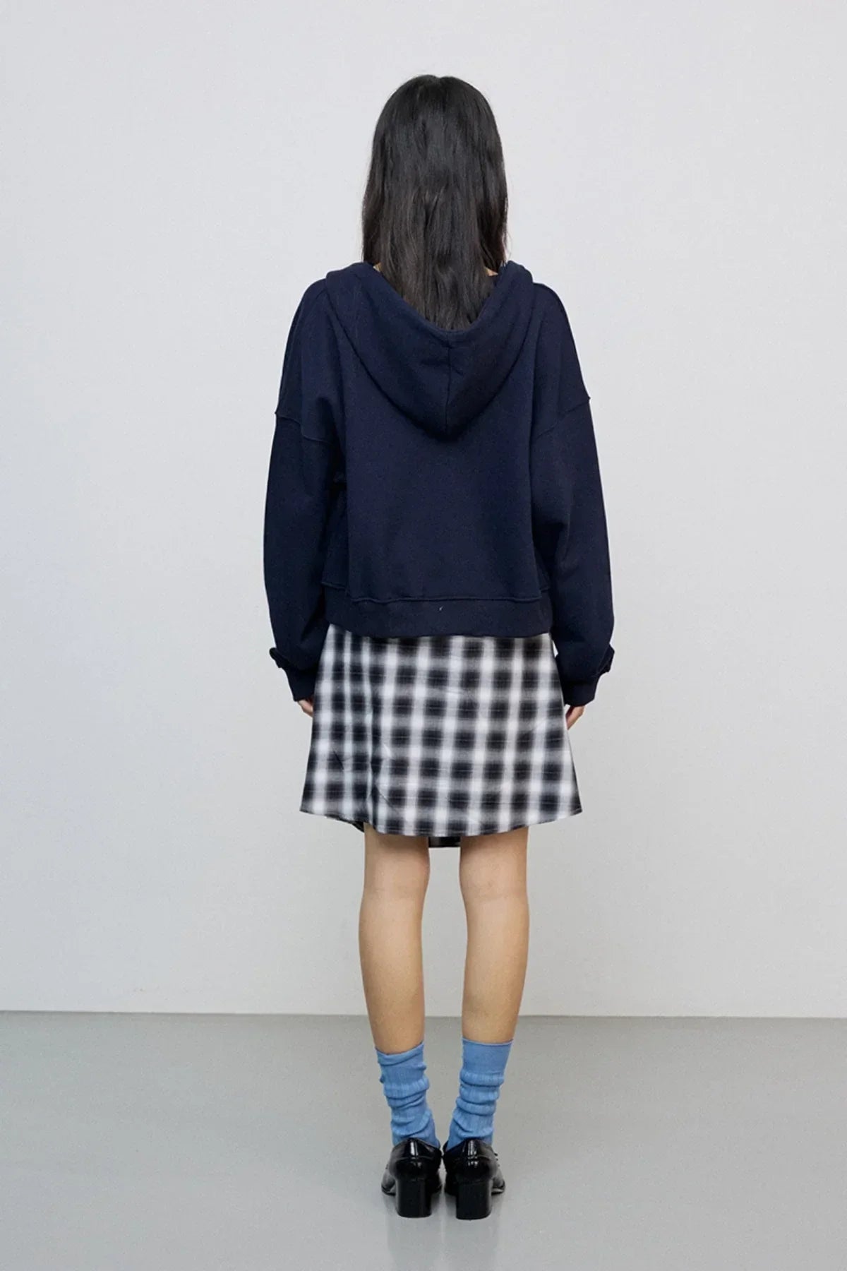Back view of woman wearing navy hoodie, black and white plaid skirt, blue socks, and black heeled shoes