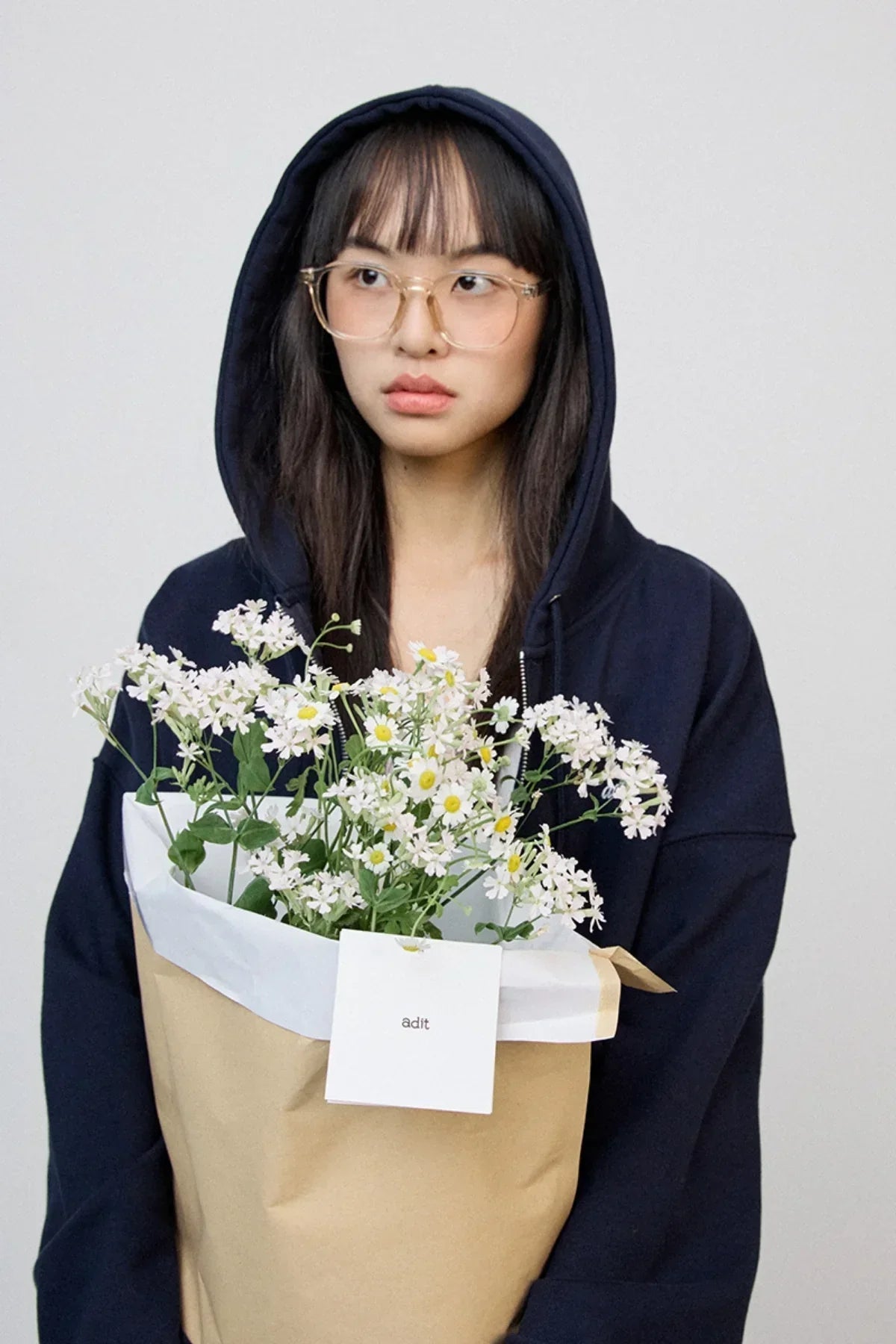 Young woman wearing navy hoodie and glasses holding a bouquet of white daisies wrapped in brown paper