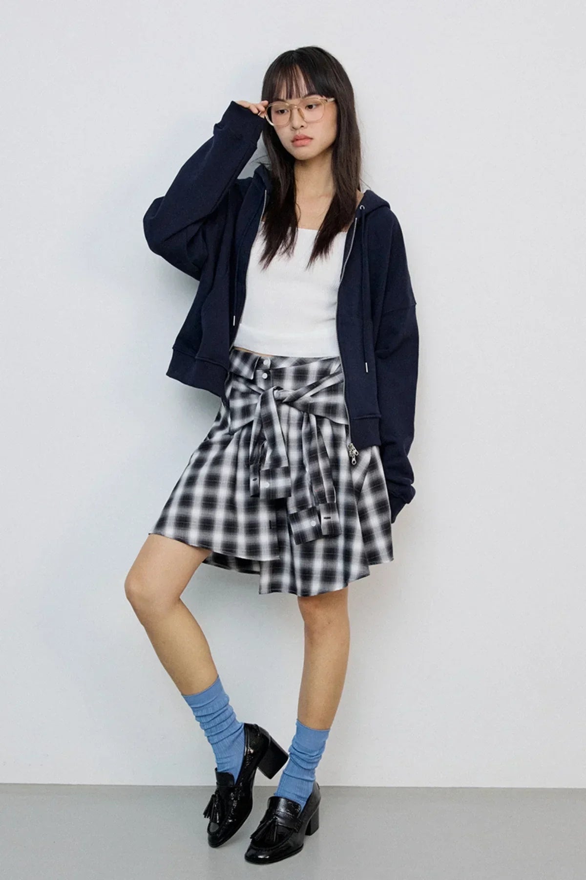 Woman wearing navy zip-up hoodie, white crop top, black and white plaid skirt, blue socks, and black loafers