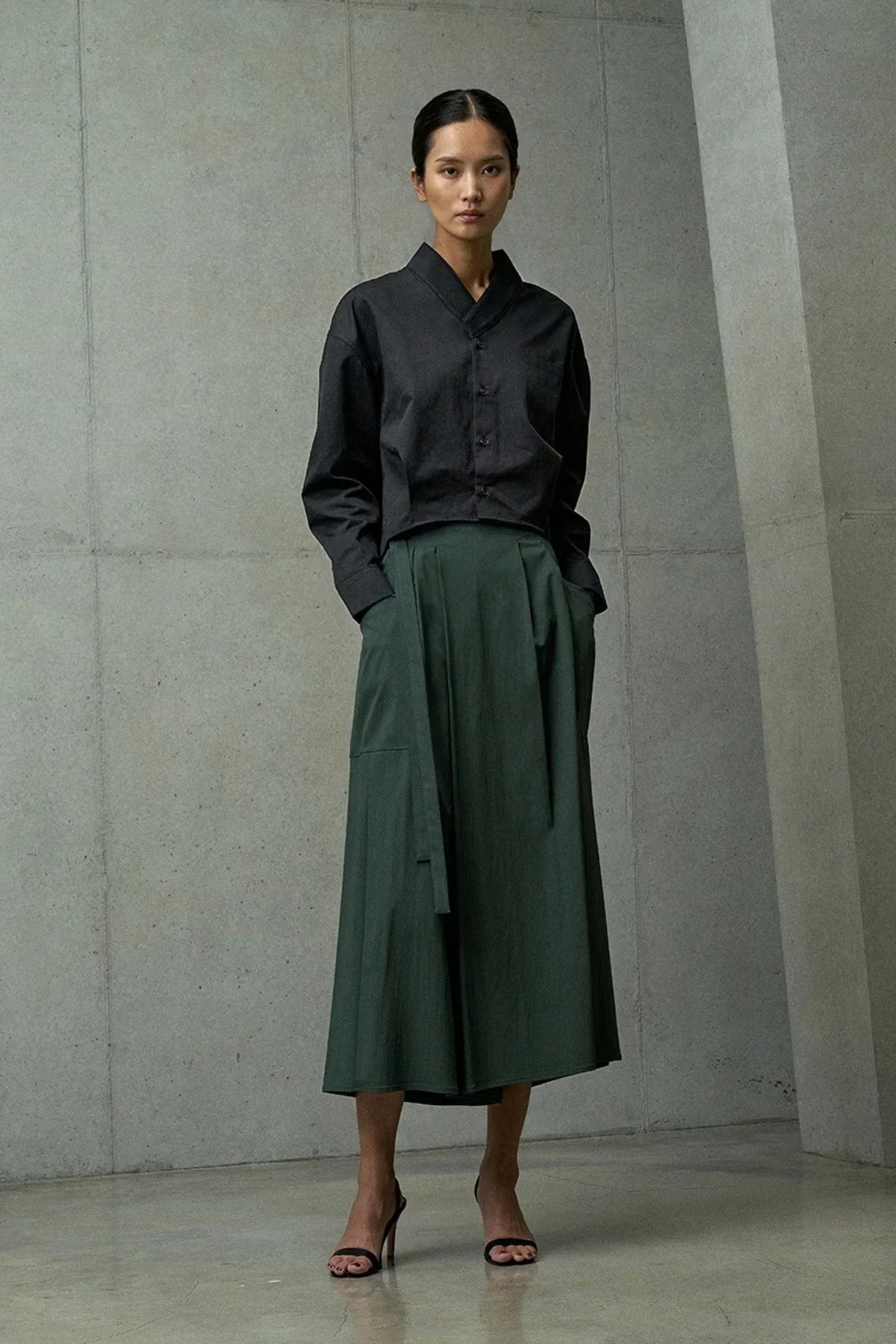 Woman wearing a black button-up cropped blouse and green midi skirt with side pocket in modern minimalist room