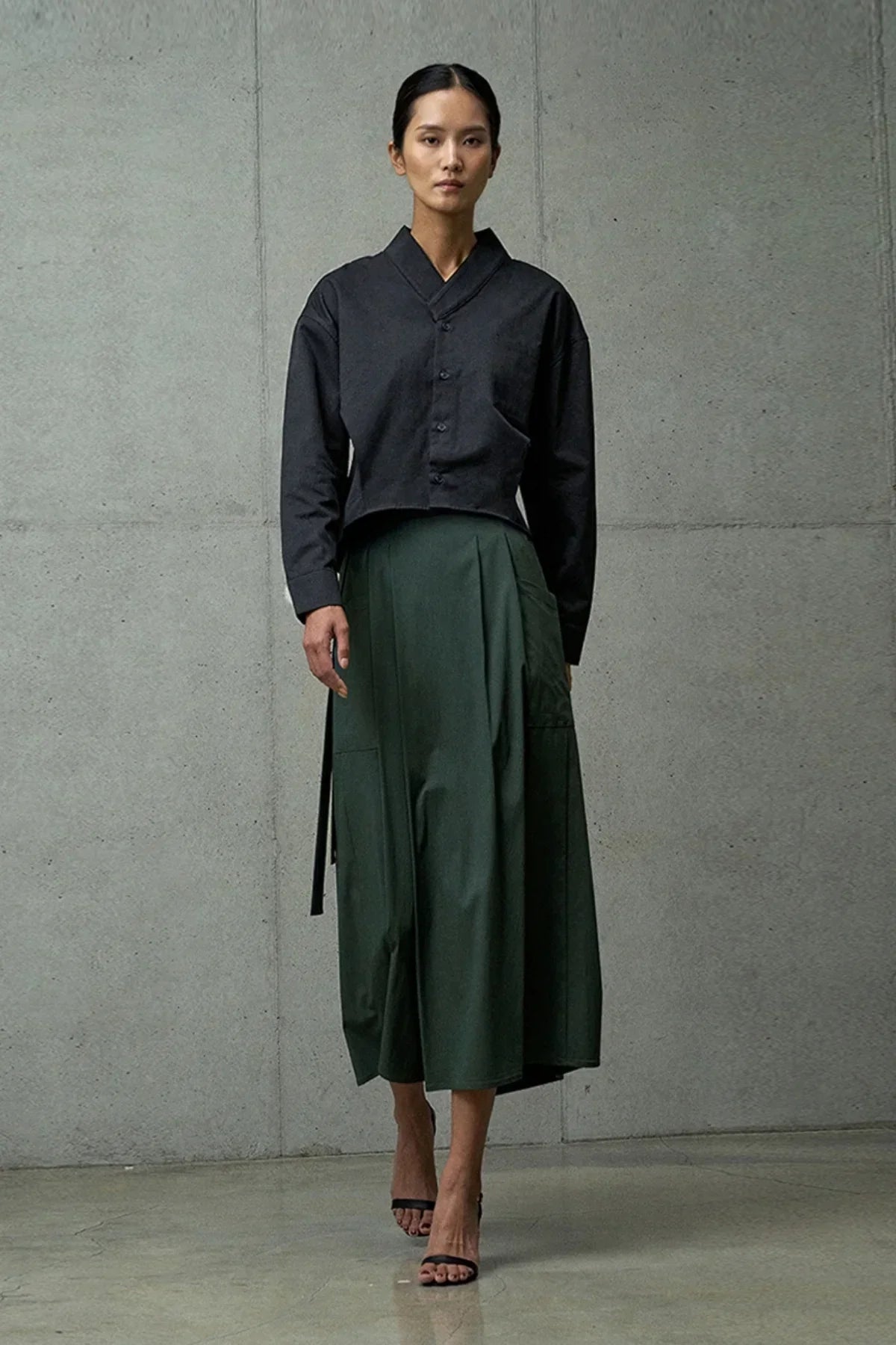 Woman wearing a black buttoned top and dark green high-waist midi skirt posing against a concrete wall