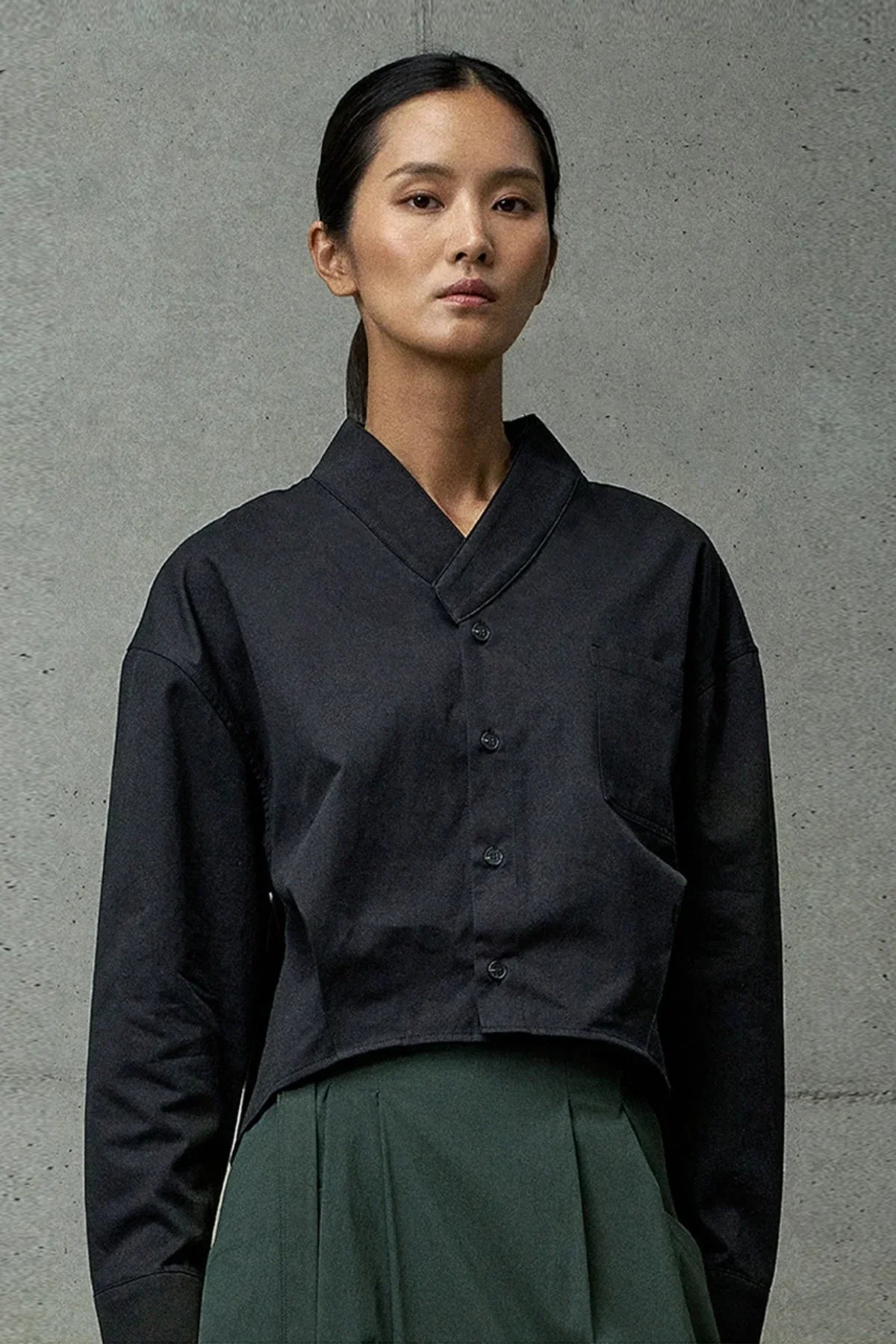 Woman in minimalist black cropped button-up shirt and high-waisted dark green pants against gray wall