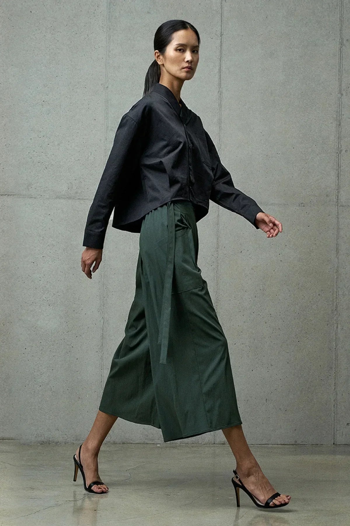 Woman walking in studio wearing black button-up top, olive wide-leg pants, and black high heel sandals