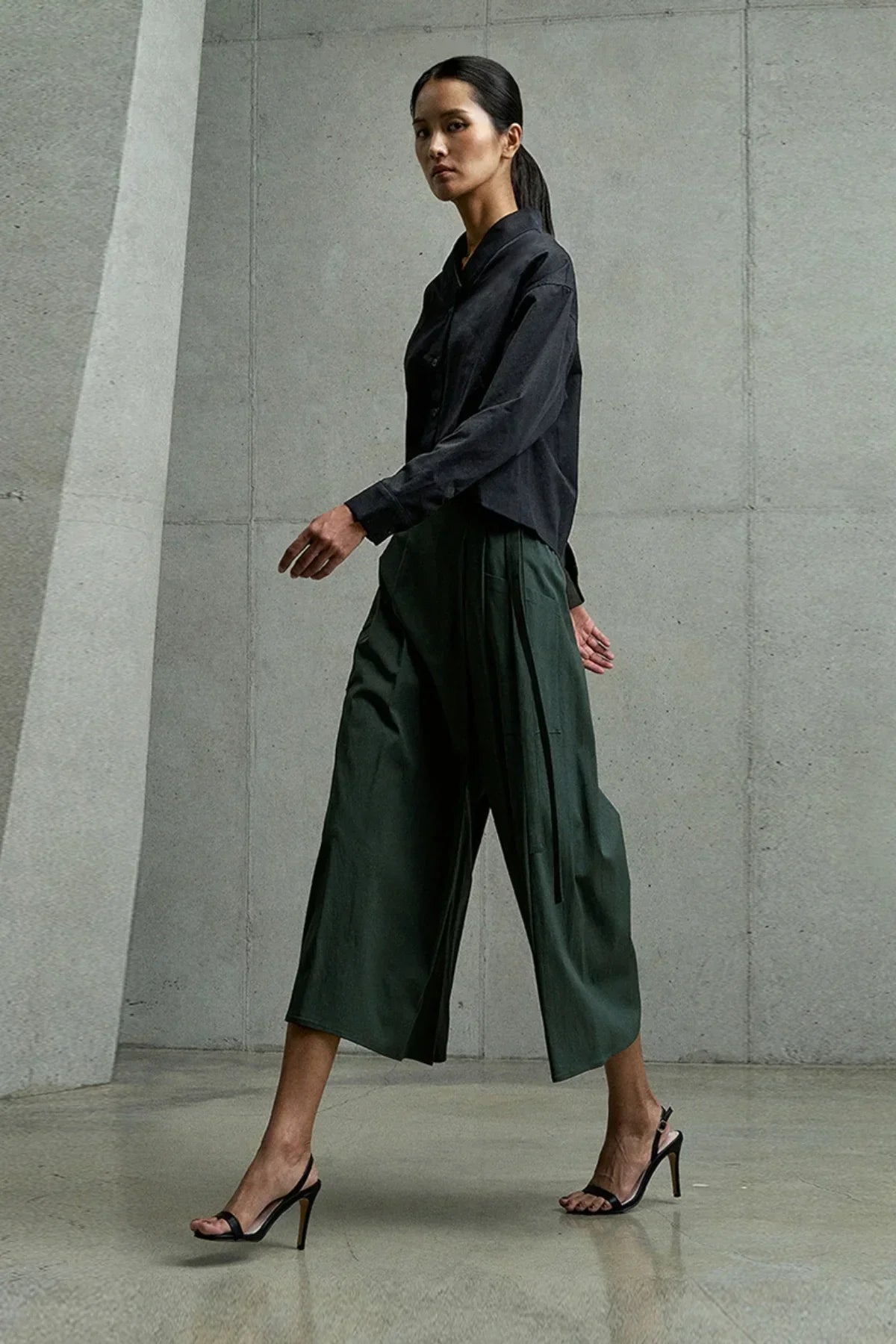 Woman walking in dark green wide-leg cropped pants with black long-sleeve shirt and black high-heeled sandals against concrete wall