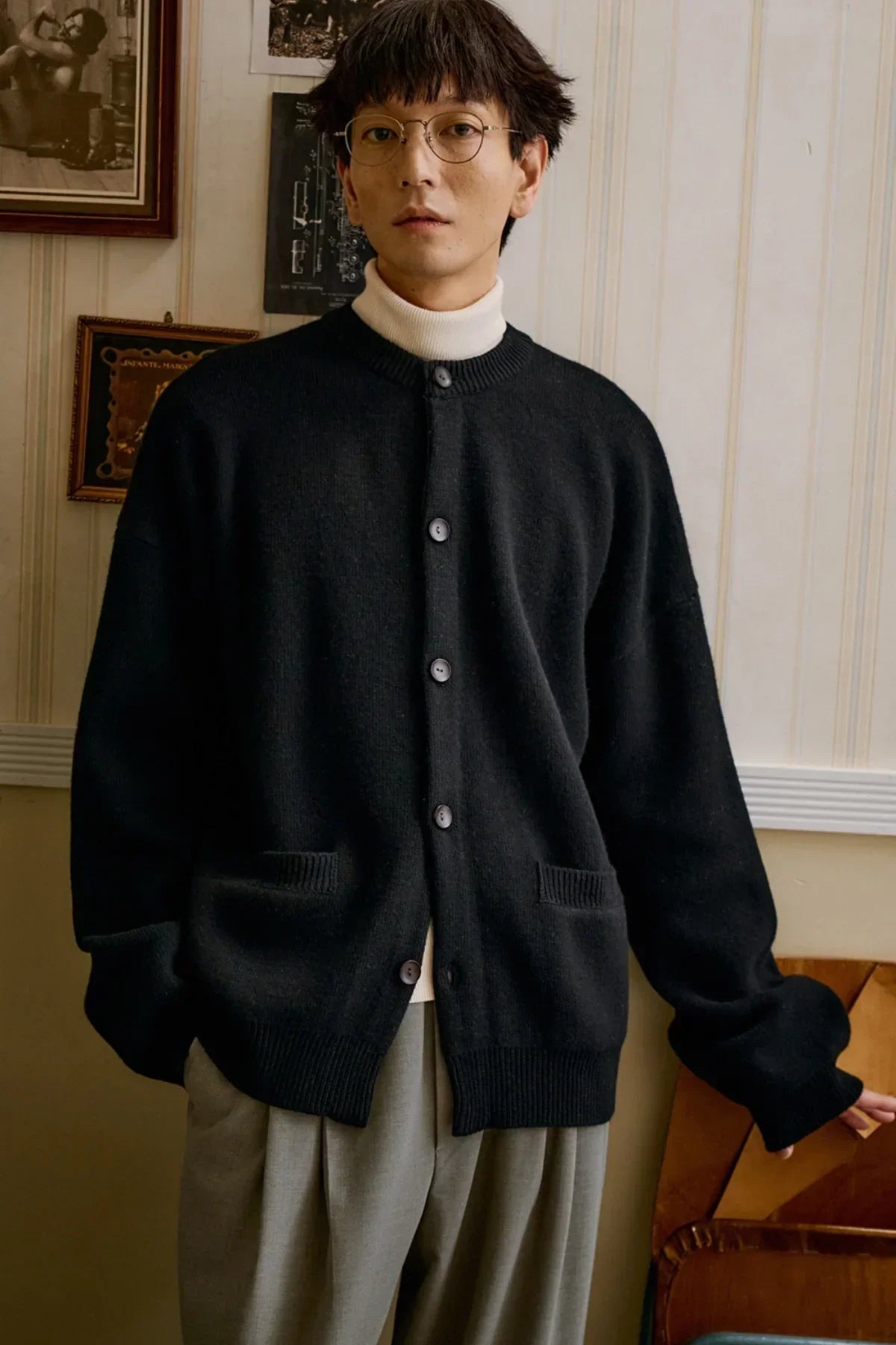Man wearing black unisex cashmere cardigan with buttons over white turtleneck and pleated pants indoors