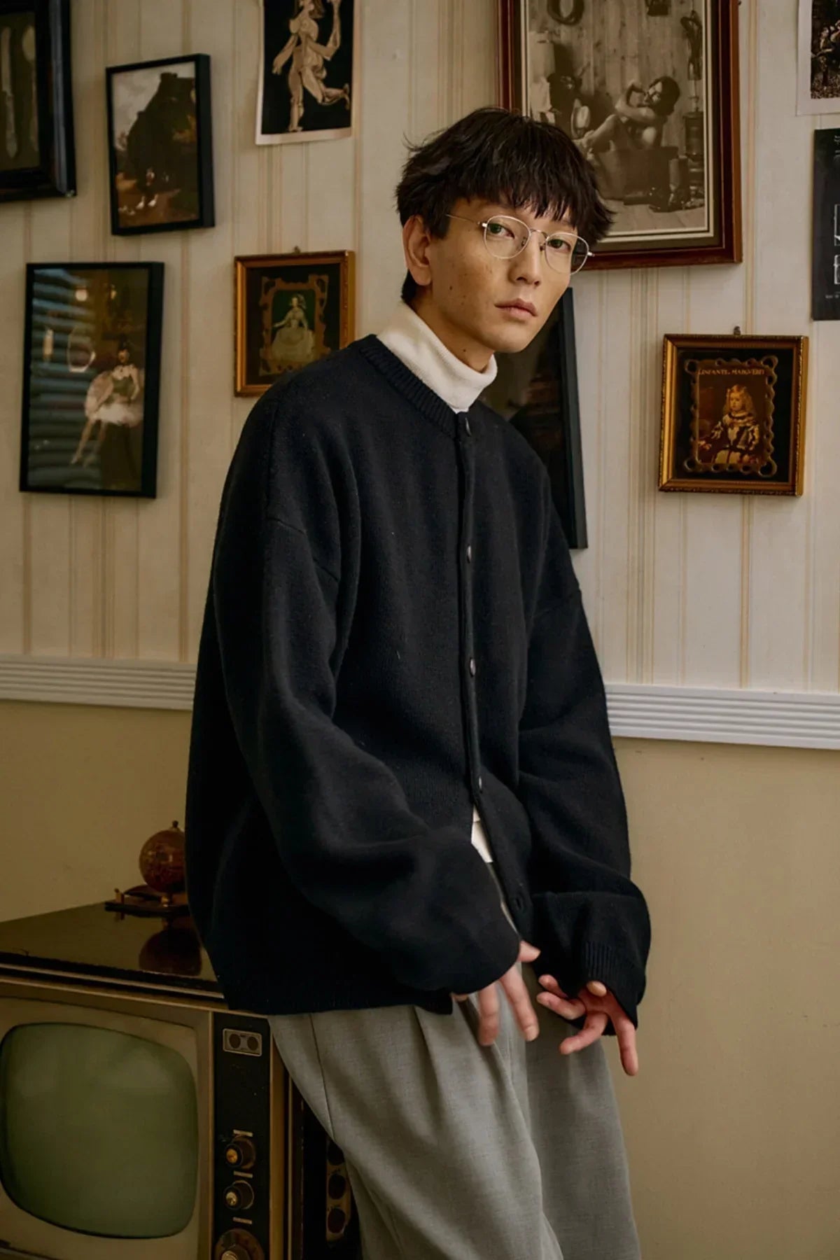 Man wearing black cashmere cardigan over white turtleneck seated indoors with framed pictures on wall