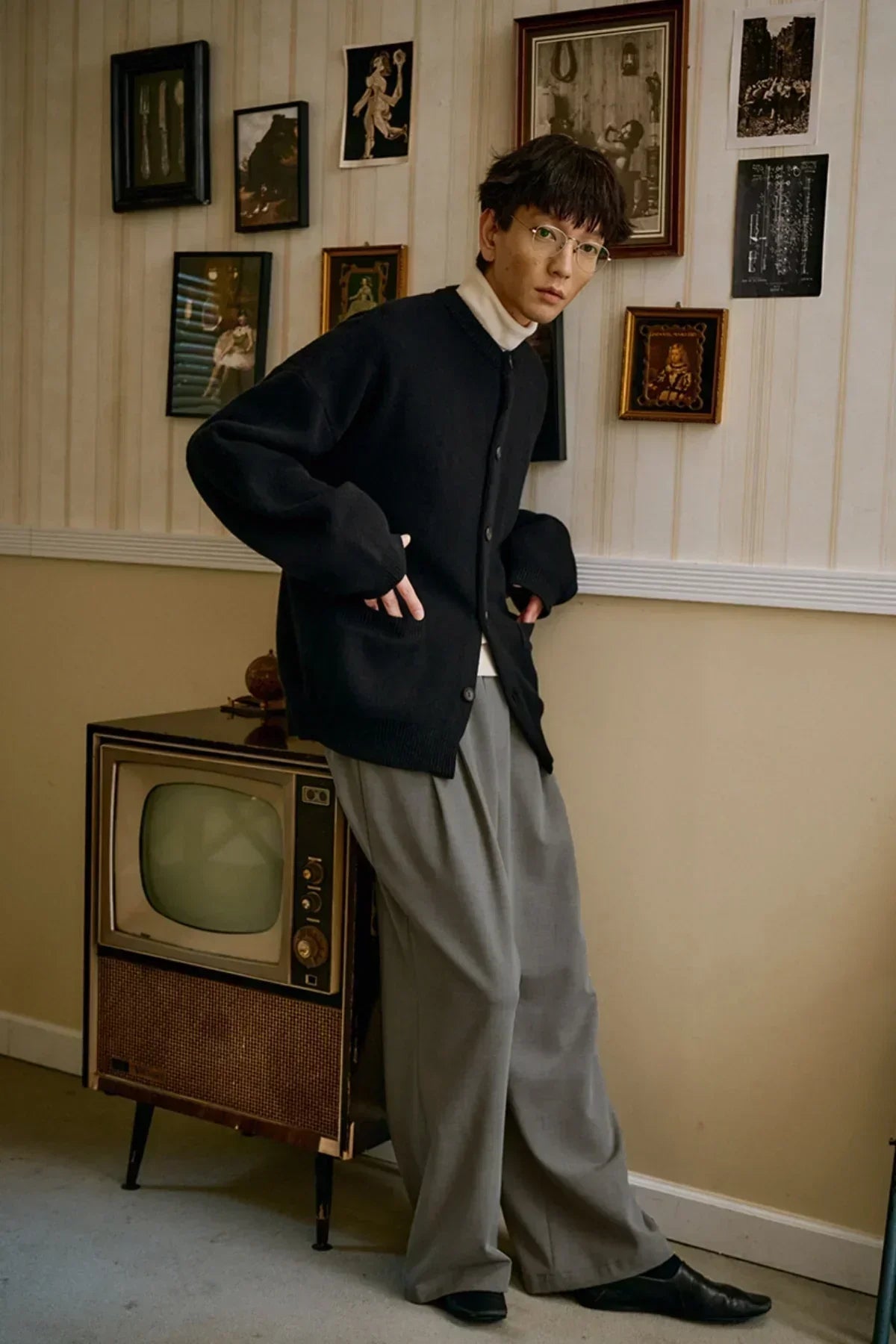 Model wearing black cashmere cardigan over white turtleneck and gray wide pants in vintage room with retro TV and framed pictures