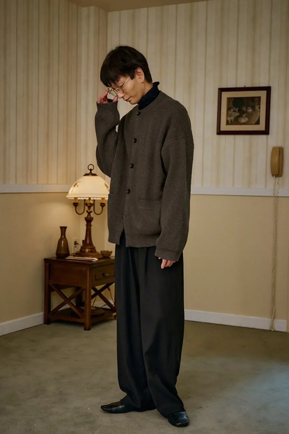 man wearing brown unisex cashmere cardigan, black wide pants, and glasses in a vintage-style room