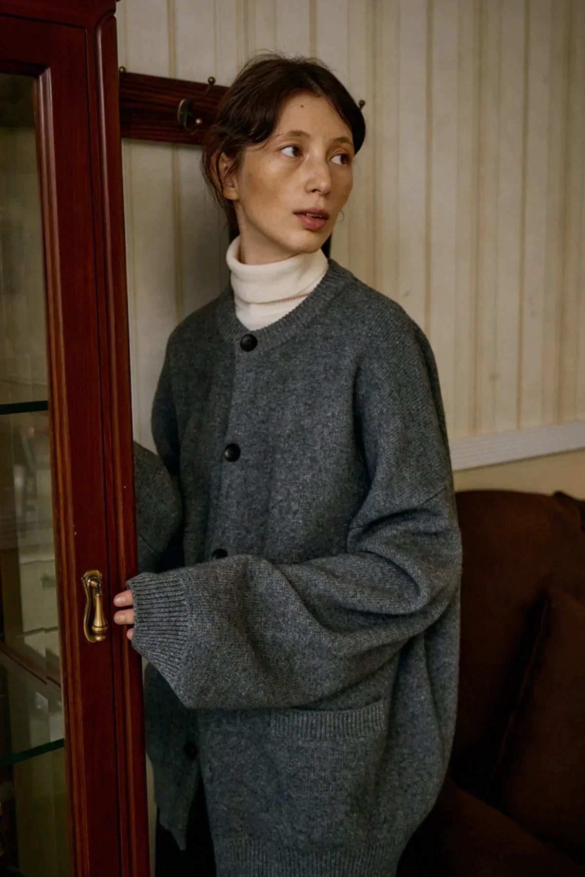 Woman wearing oversized gray cashmere cardigan and white turtleneck indoors near wooden cabinet
