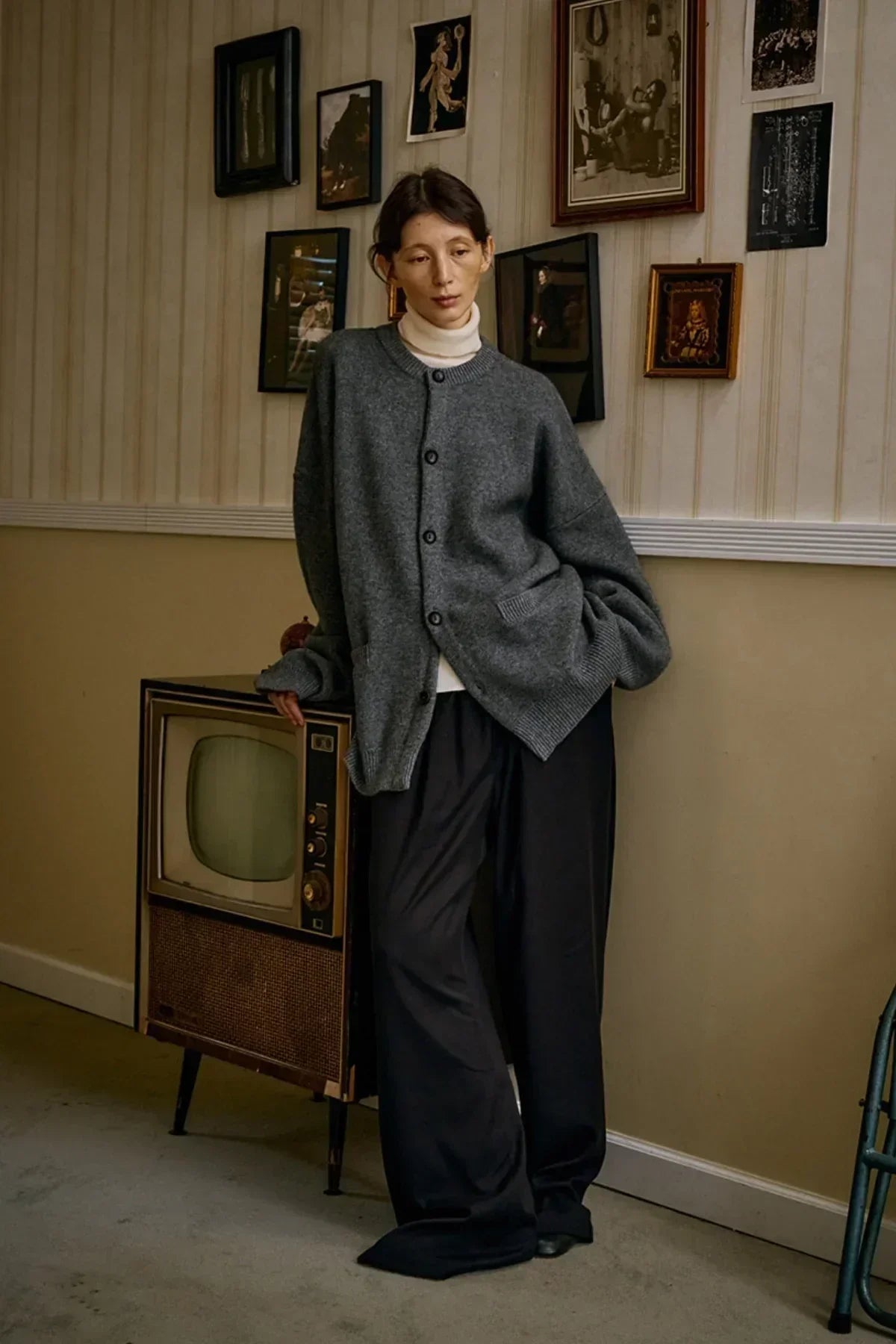Woman wearing gray oversized cashmere cardigan and black wide-leg pants leaning on vintage TV in cozy room