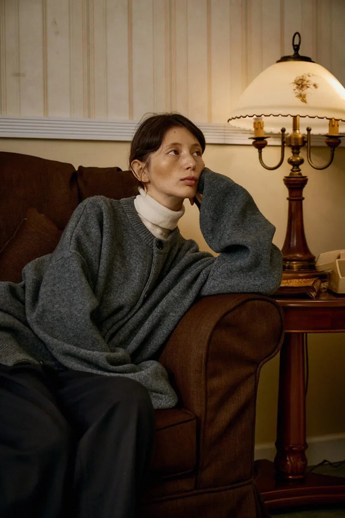 Woman lounging on brown sofa wearing gray cashmere cardigan and beige turtleneck in cozy vintage living room