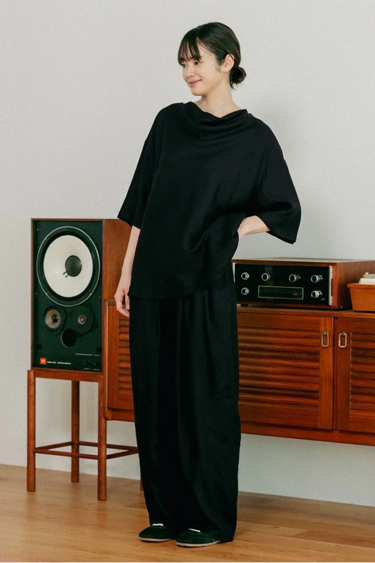 Woman wearing black unisex cowl neck t-shirt and wide black pants indoors with retro speaker and wooden cabinet