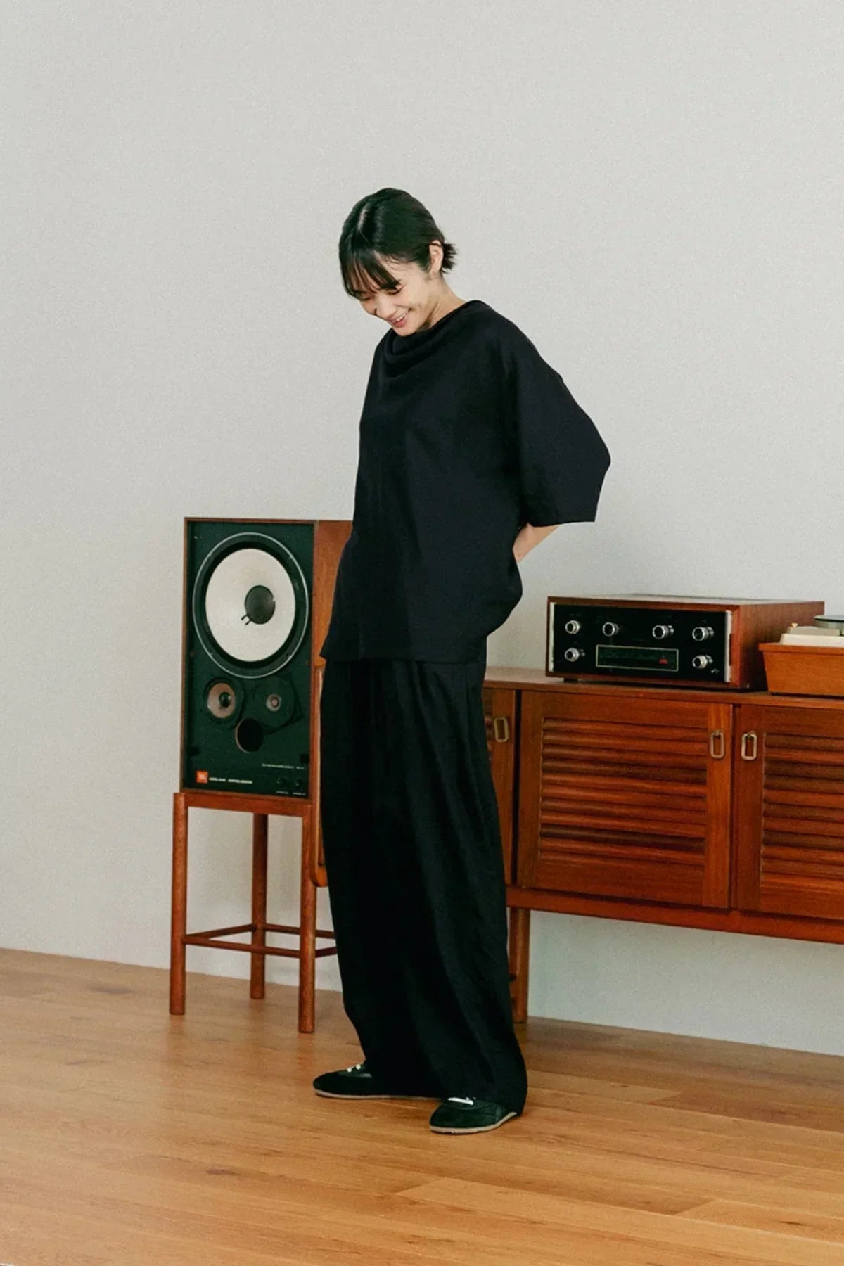 Person wearing loose black cowl neck t-shirt and wide pants standing in room with wooden speaker and console
