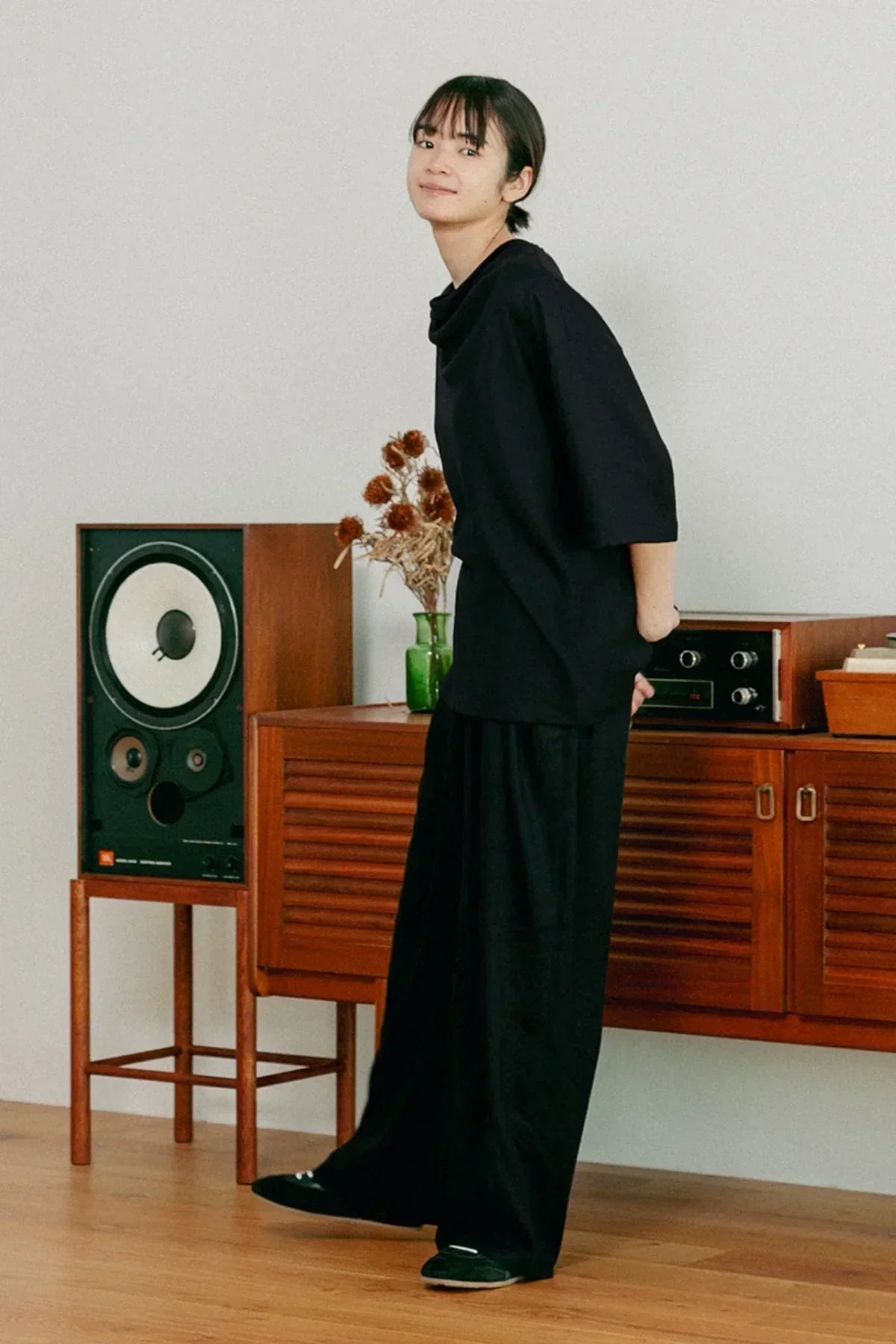 Woman in black unisex cowl neck t-shirt and wide pants standing in modern room with vintage wooden speaker and sideboard