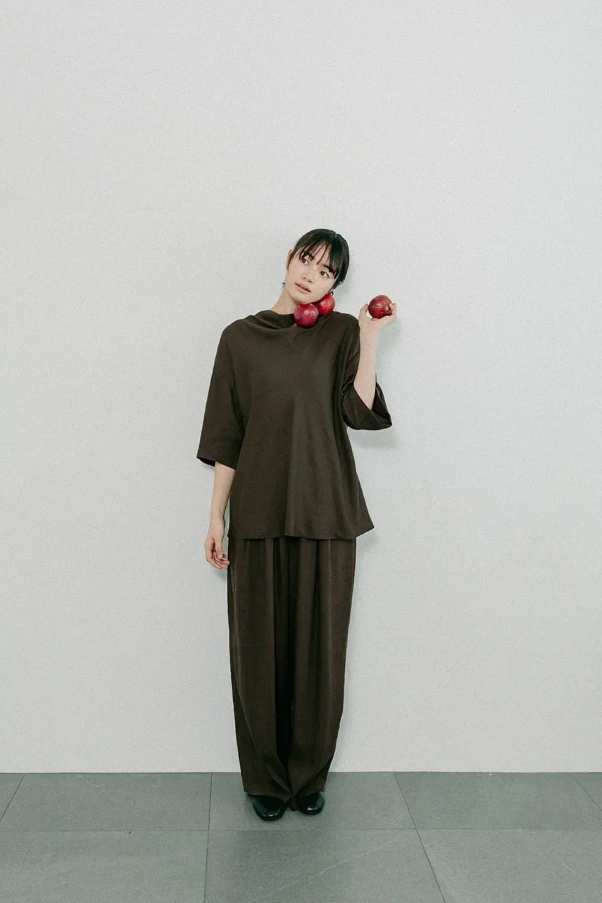 Woman wearing brown unisex cowl neck t-shirt and wide pants holding red apples against plain wall