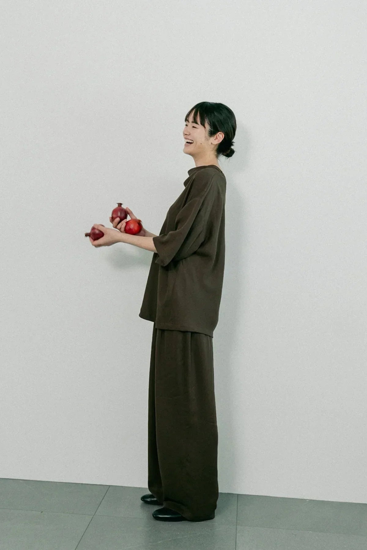 Smiling woman in brown unisex cowl neck t-shirt and wide pants holding red onions against white wall