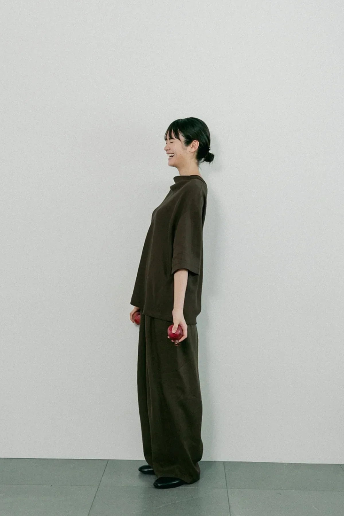 Smiling woman in brown unisex cowl neck t-shirt and wide pants holding red apples, standing against white wall