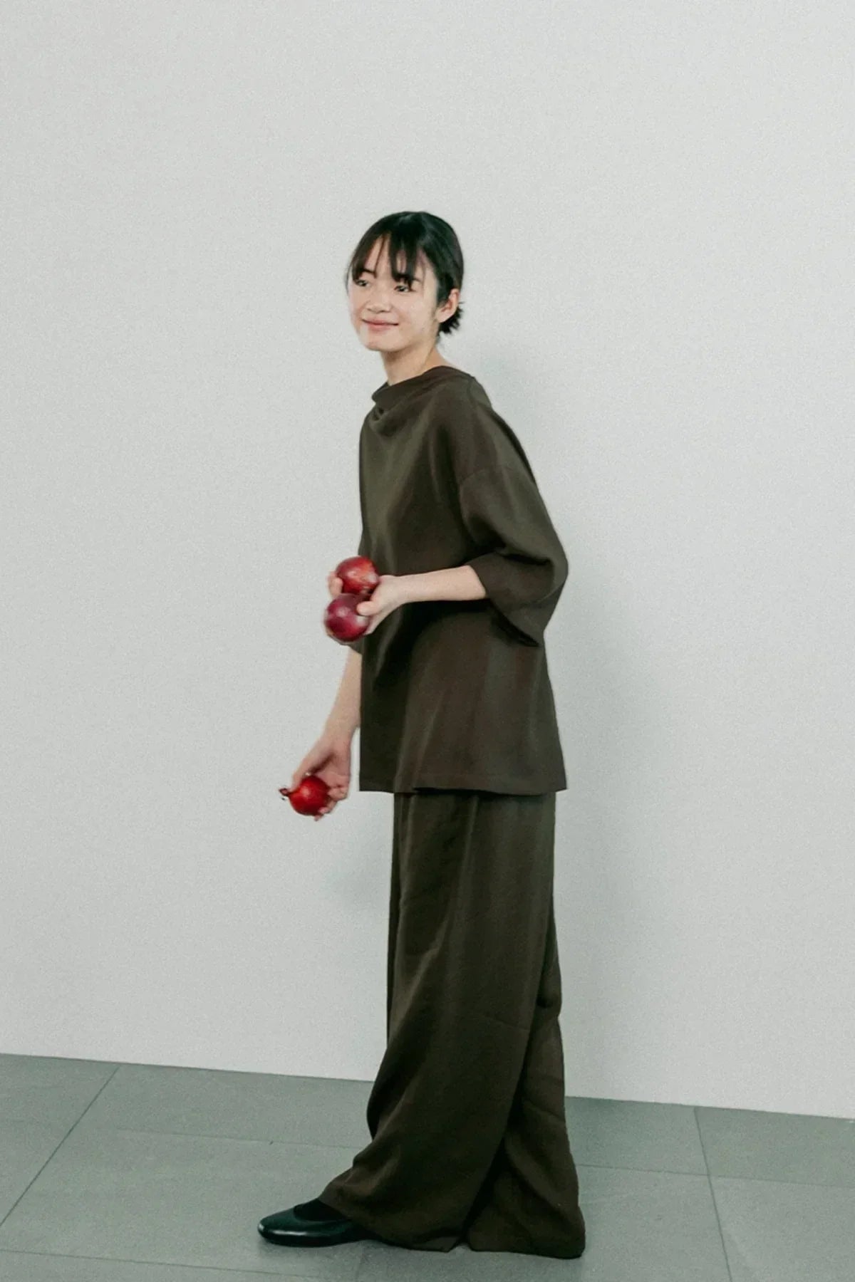 Woman wearing brown unisex cowl neck t-shirt and wide pants holding red onions against plain background
