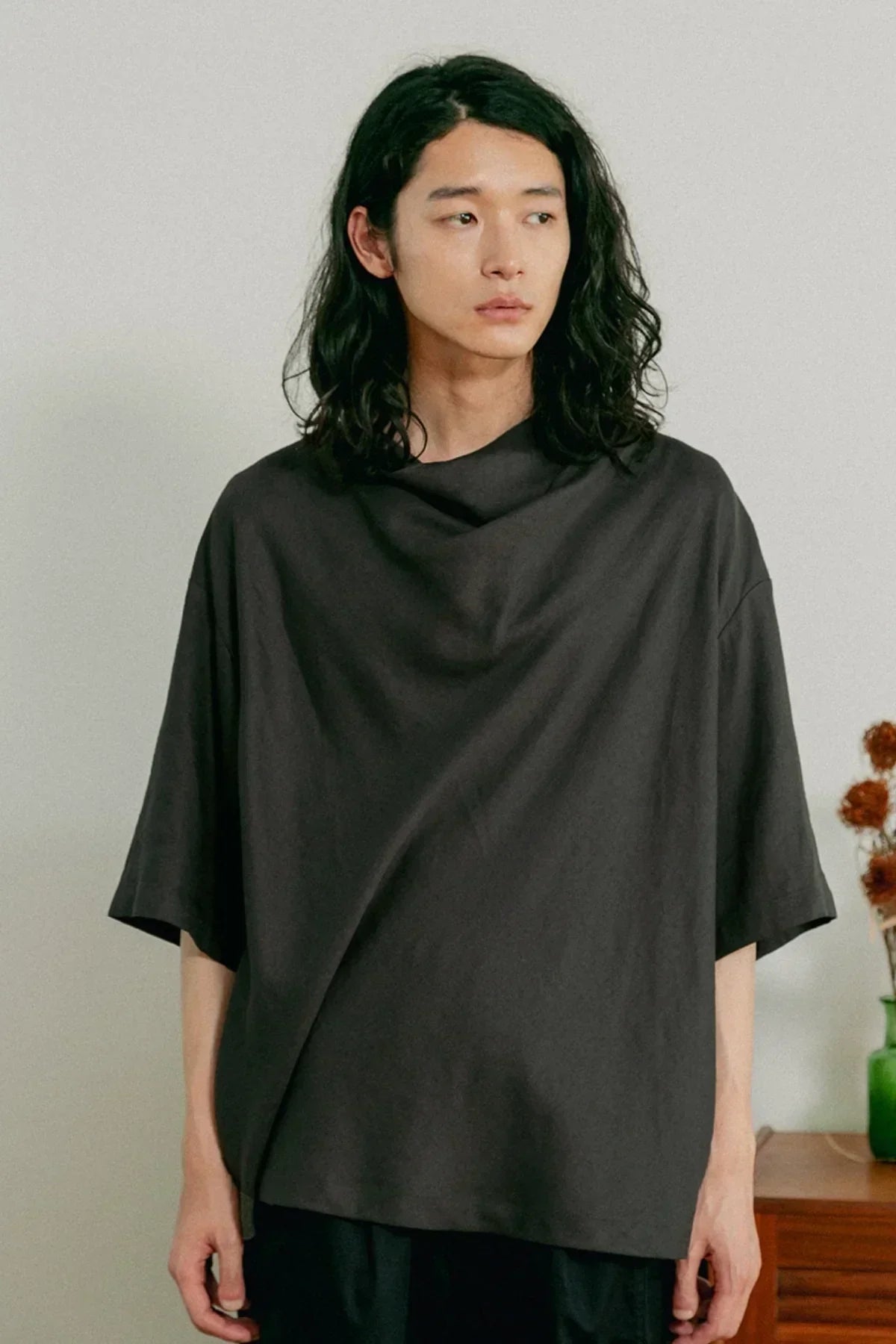 Young man with long black hair wearing a loose dark gray unisex cowl neck t-shirt indoors