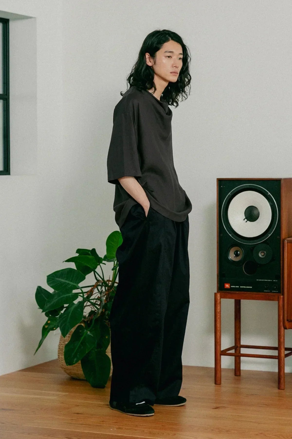 person wearing loose black pants and dark gray cowl neck t-shirt standing indoors by speaker and plant