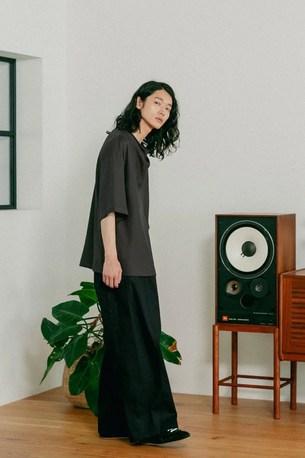 Young person in loose black t-shirt and wide black pants standing indoors near vintage speaker and green plant
