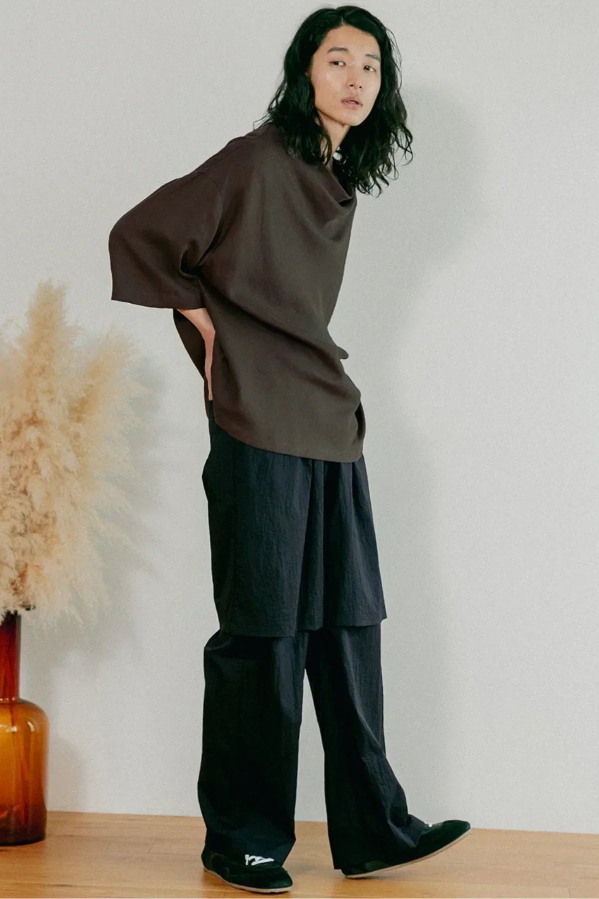 Model wearing oversized brown cowl neck top and loose black pants in minimalist studio with pampas grass