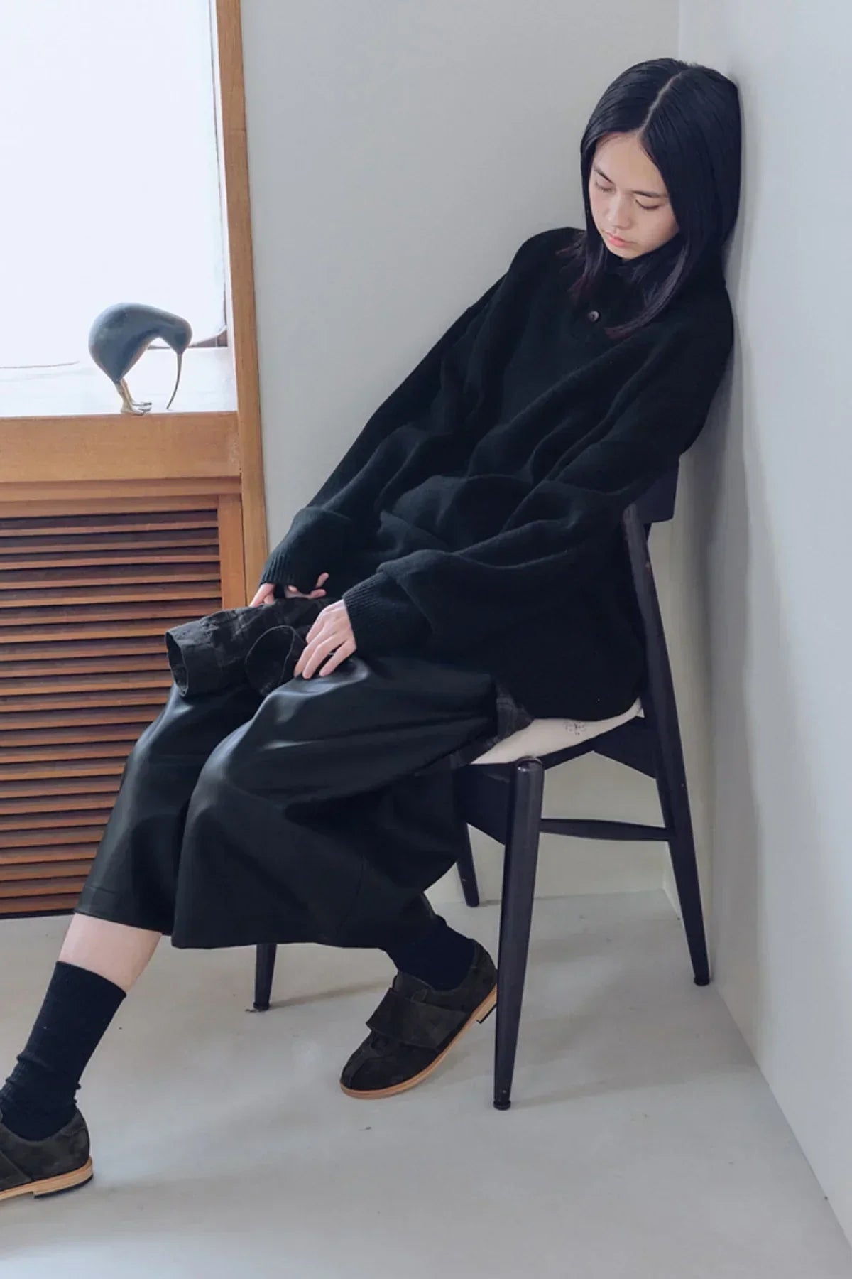 Young woman in black knit Henley neck sweater and wide black pants sitting on chair by wall