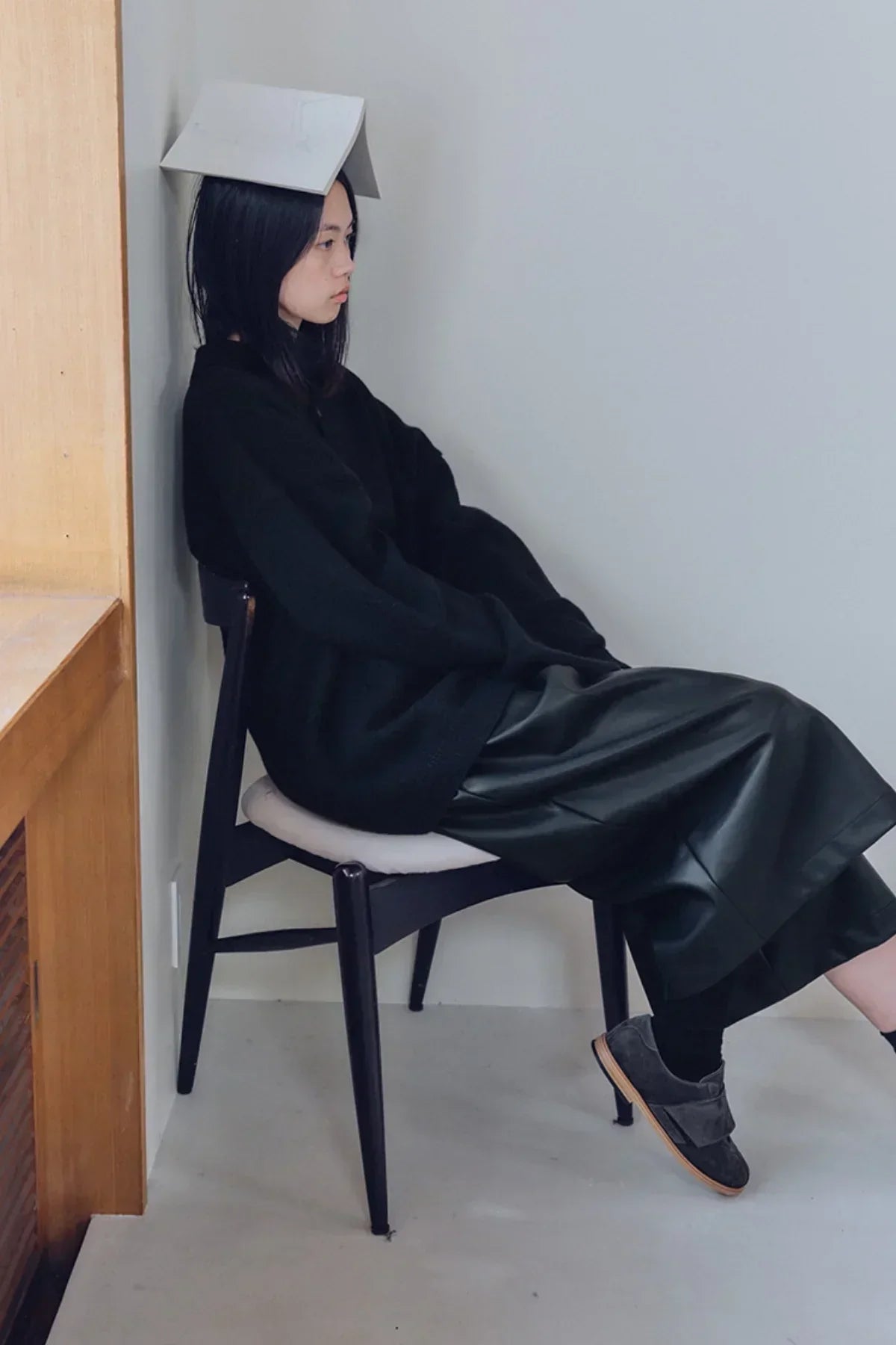 Woman sitting sideways on black chair against gray wall wearing black knit Henley neck shirt and leather skirt