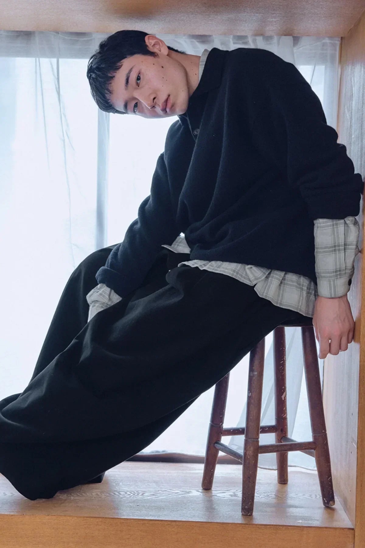 Young man wearing black knit Henley shirt over plaid shirt, sitting on wooden stool in bright room