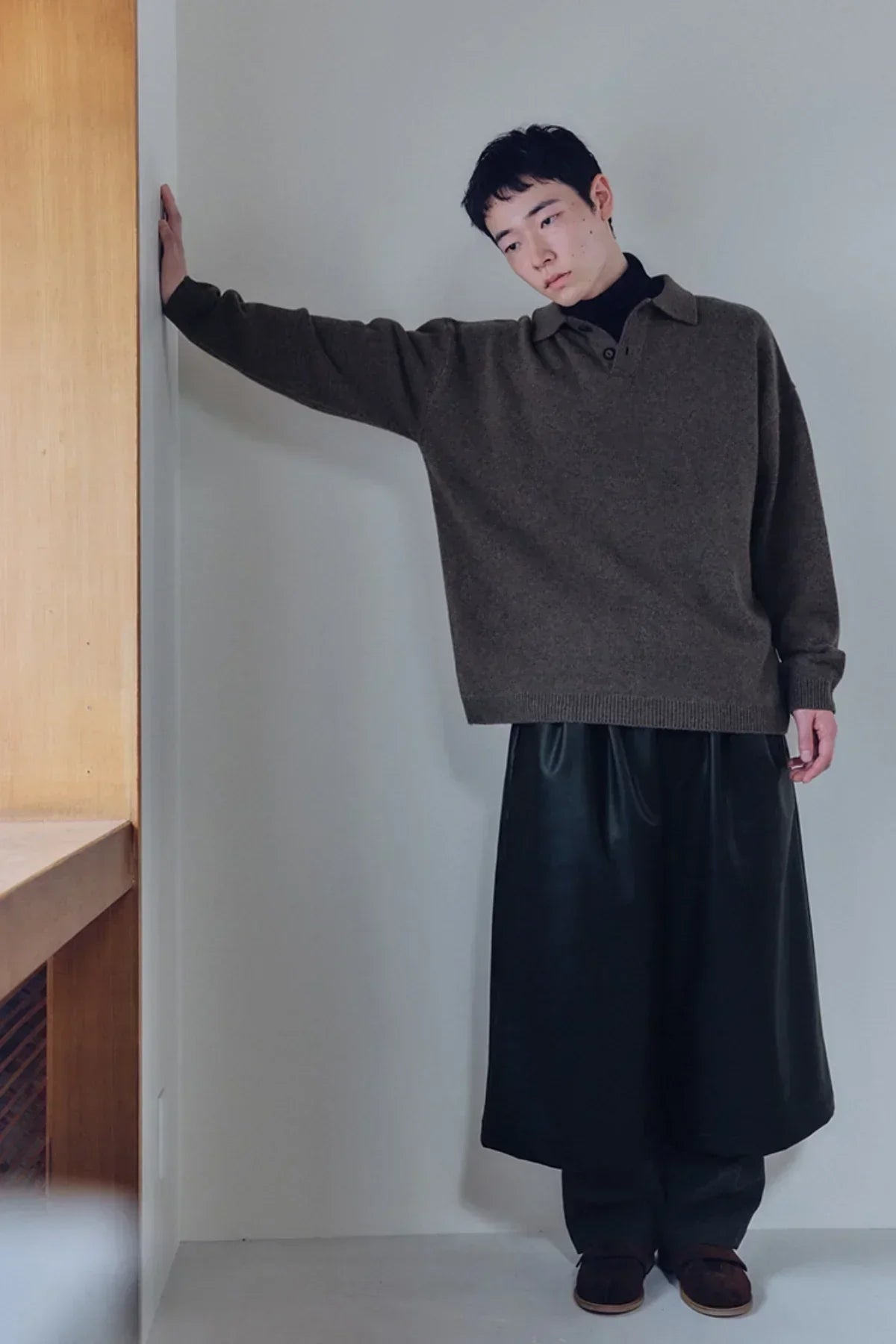 Young man wearing brown knit Henley neck sweater and loose black skirt pants against white wall