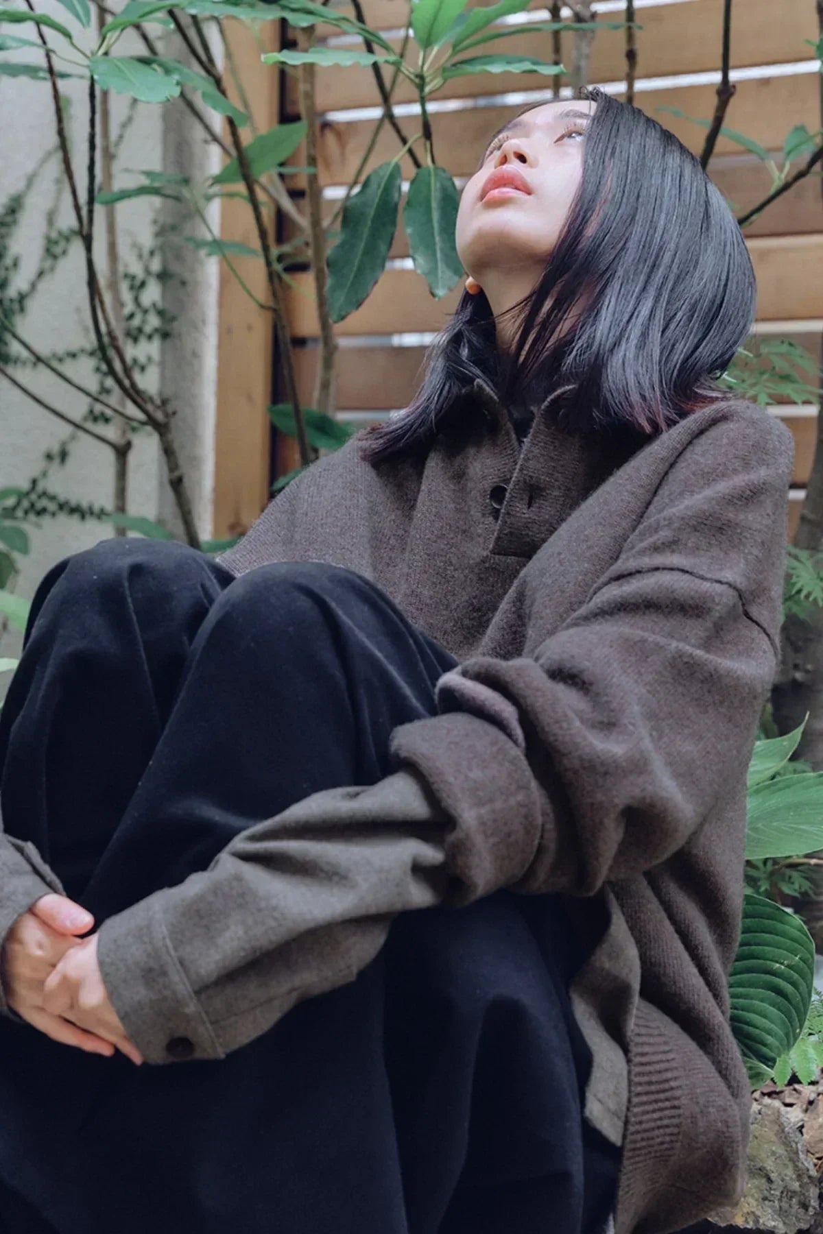 Person wearing a brown knit Henley neck sweater sitting outdoors against wooden fence and plants