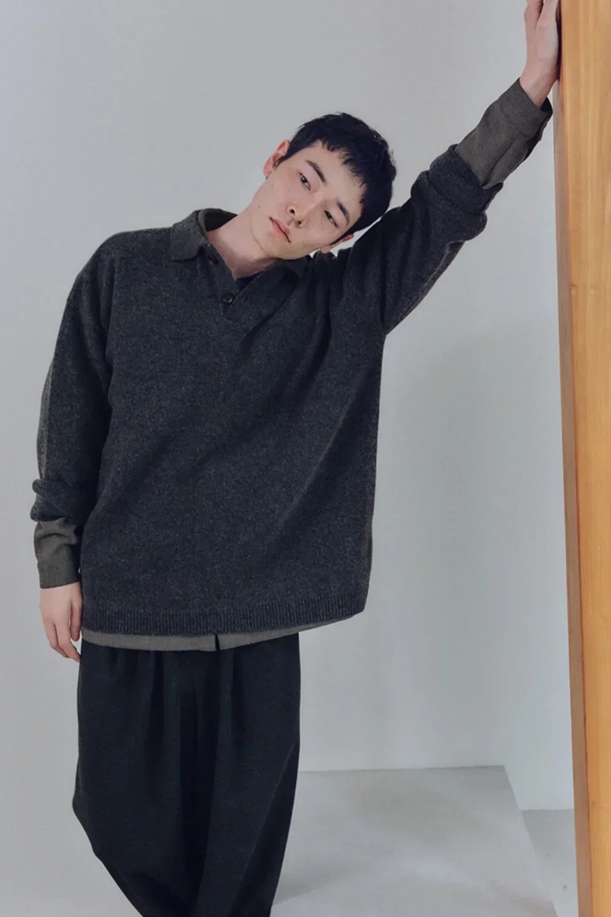 Man wearing dark charcoal unisex knit Henley neck shirt with long sleeves standing by wooden frame