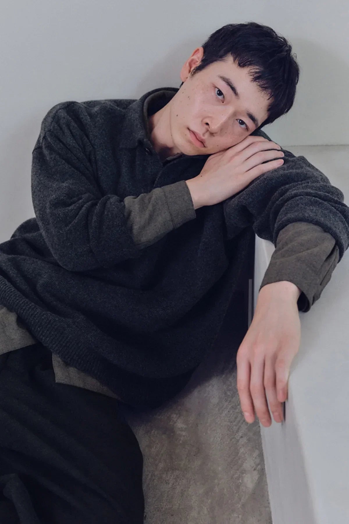 Man wearing charcoal gray unisex knit henley neck shirt leaning on white surface in studio